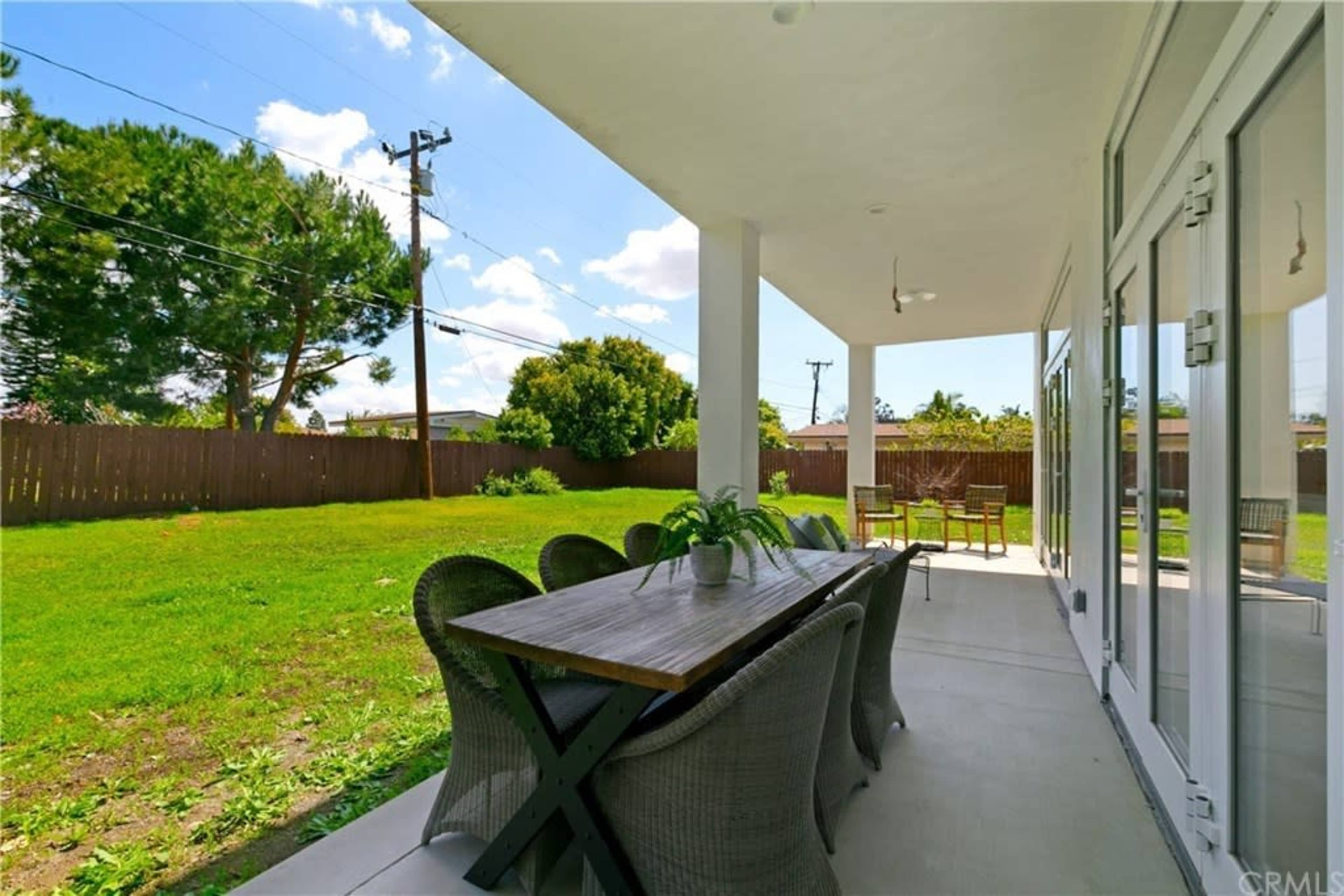 A spacious patio features a long dining table surrounded by chairs and overlooks a grassy backyard with trees.