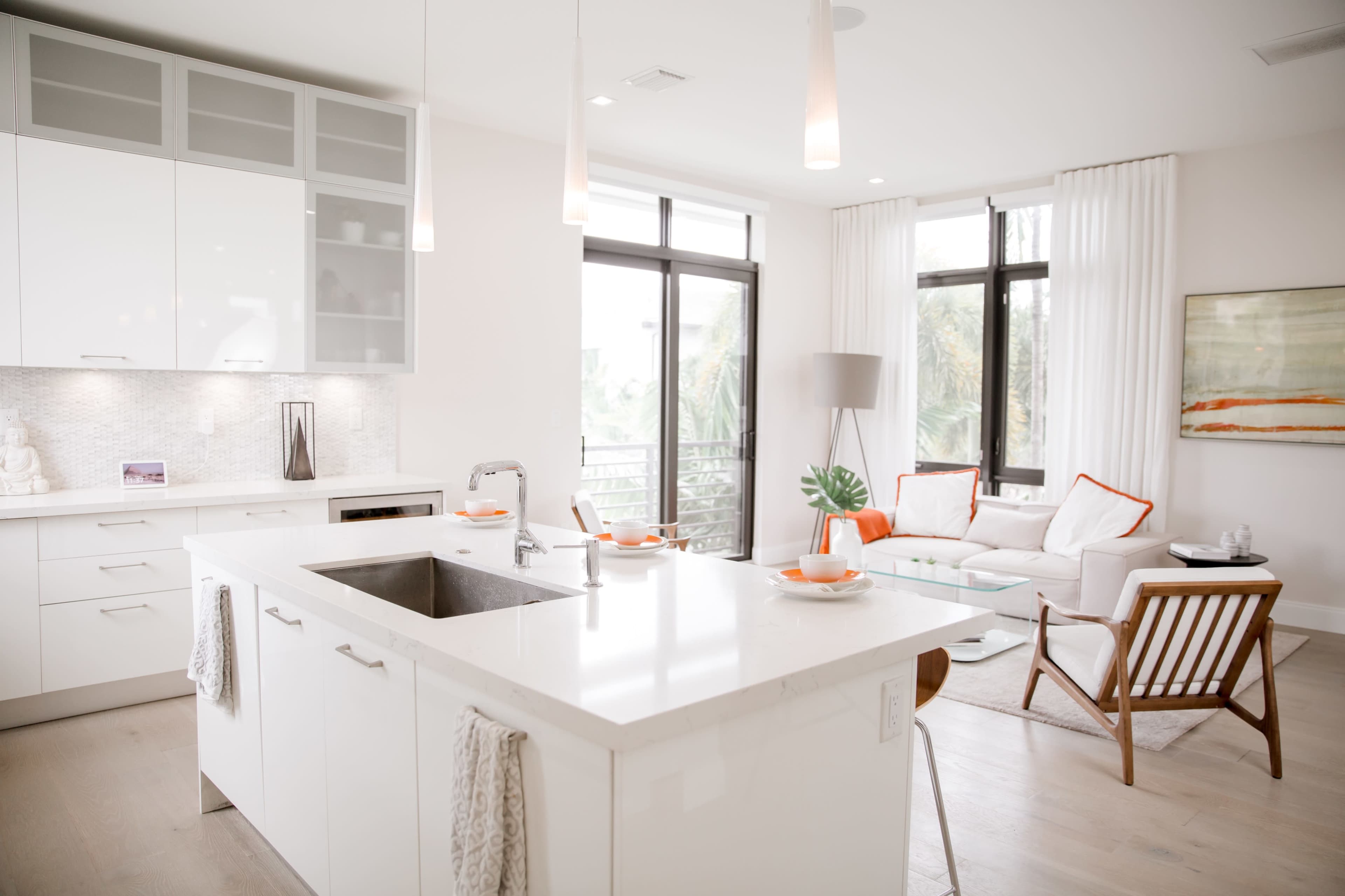 A modern kitchen with white cabinetry and an island is connected to a bright, open living area featuring a couch and armchair with orange accents.
