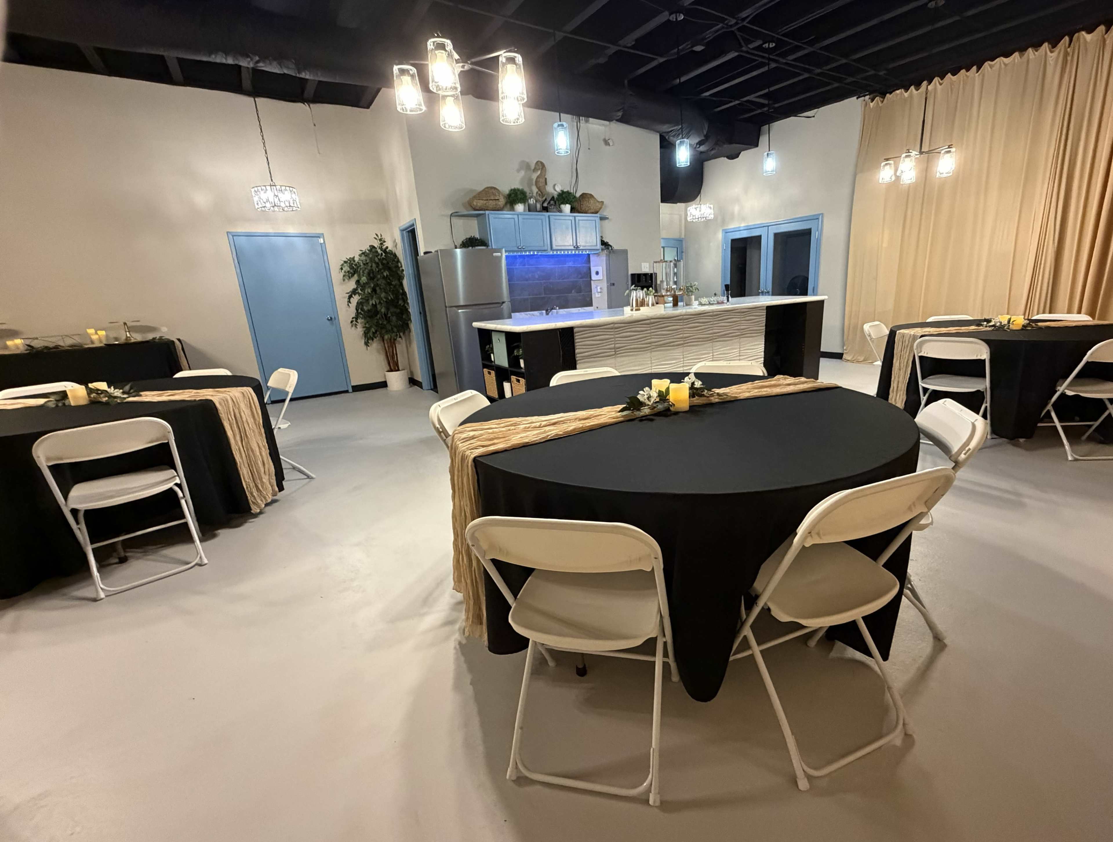 The image shows a spacious venue with several round tables covered in black tablecloths, each adorned with a centerpiece, and a kitchen area visible in the background.