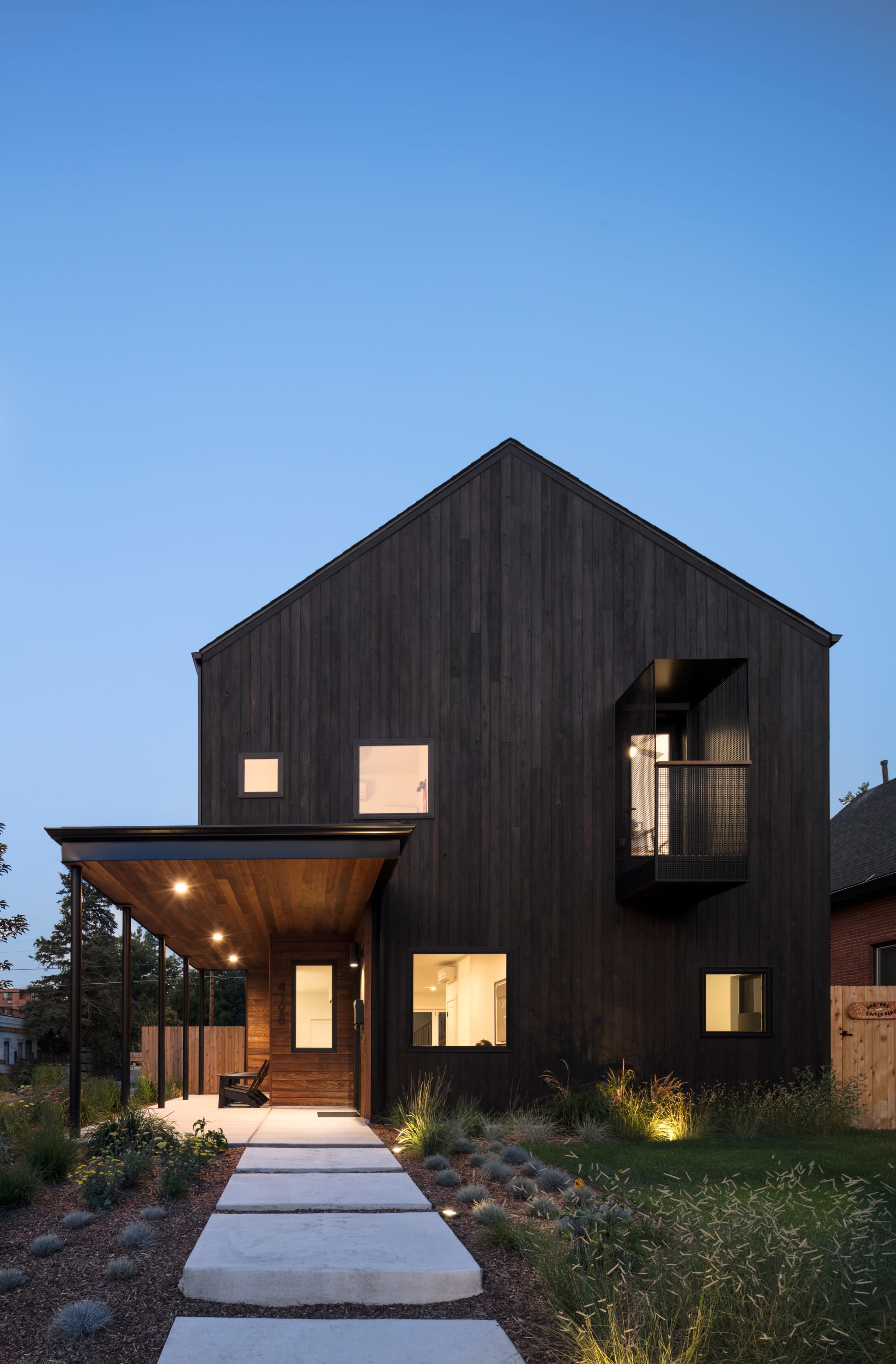 The image shows a modern black wooden house with large windows and a covered porch, set against a twilight sky.