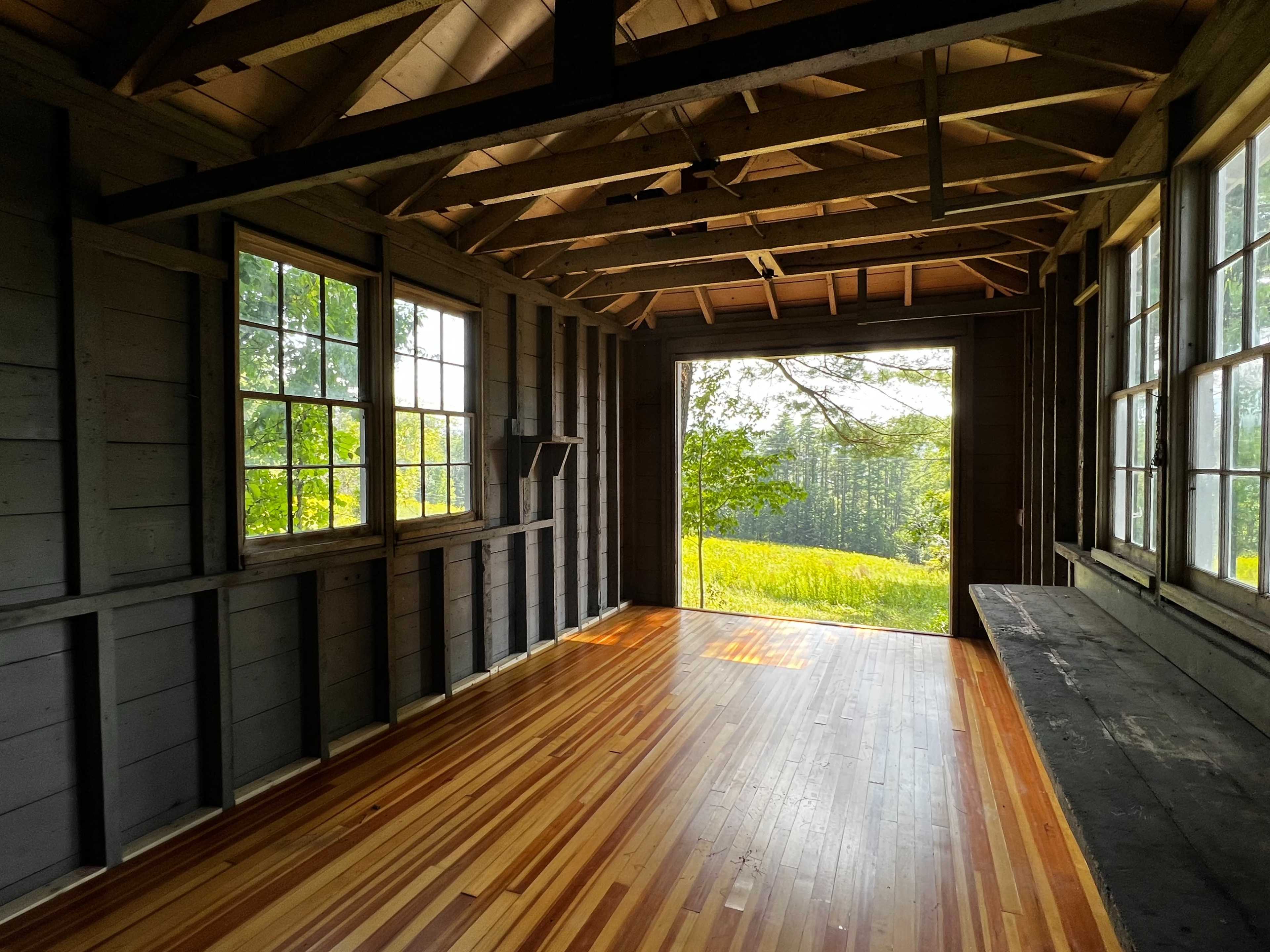 The interior of a wooden cabin features large windows that open to a view of a grassy field and trees outside, with polished wooden flooring.