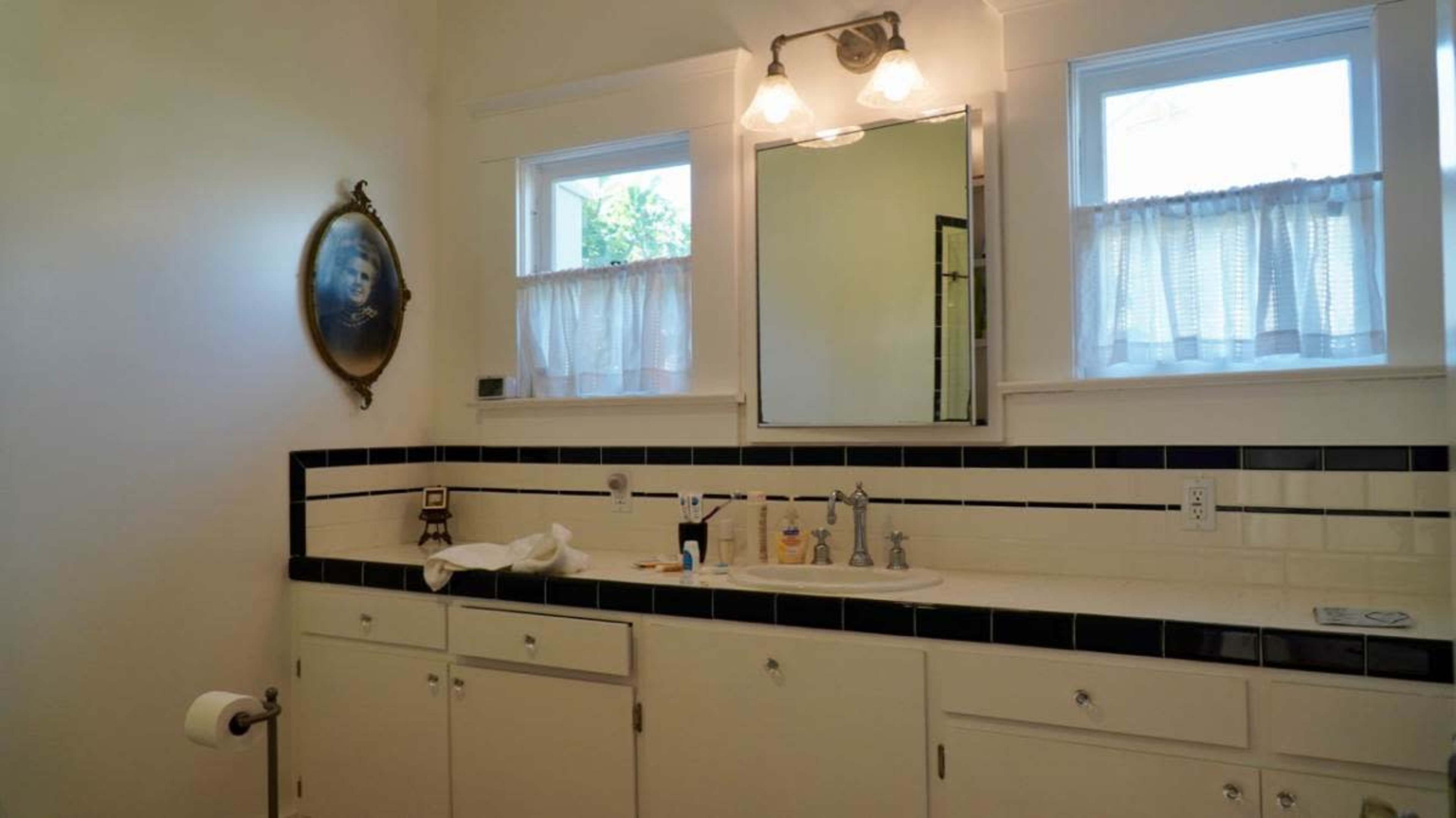 The image shows a bathroom with white walls, a mirror above the sink, and two windows with sheer curtains, featuring a vintage portrait on the wall.