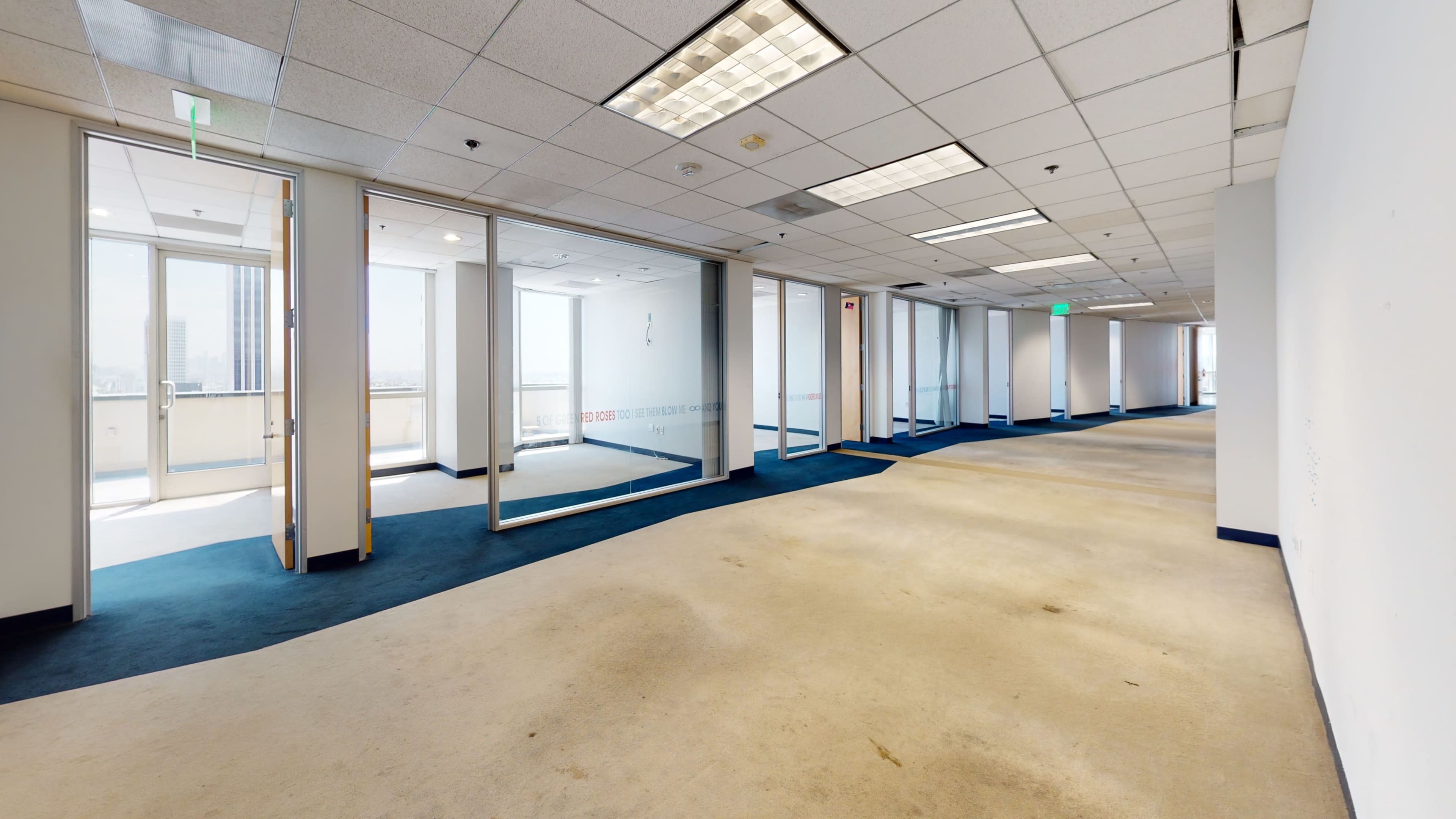 A corridor in an office building features large glass windows on either side and a carpeted floor, leading to several empty rooms.