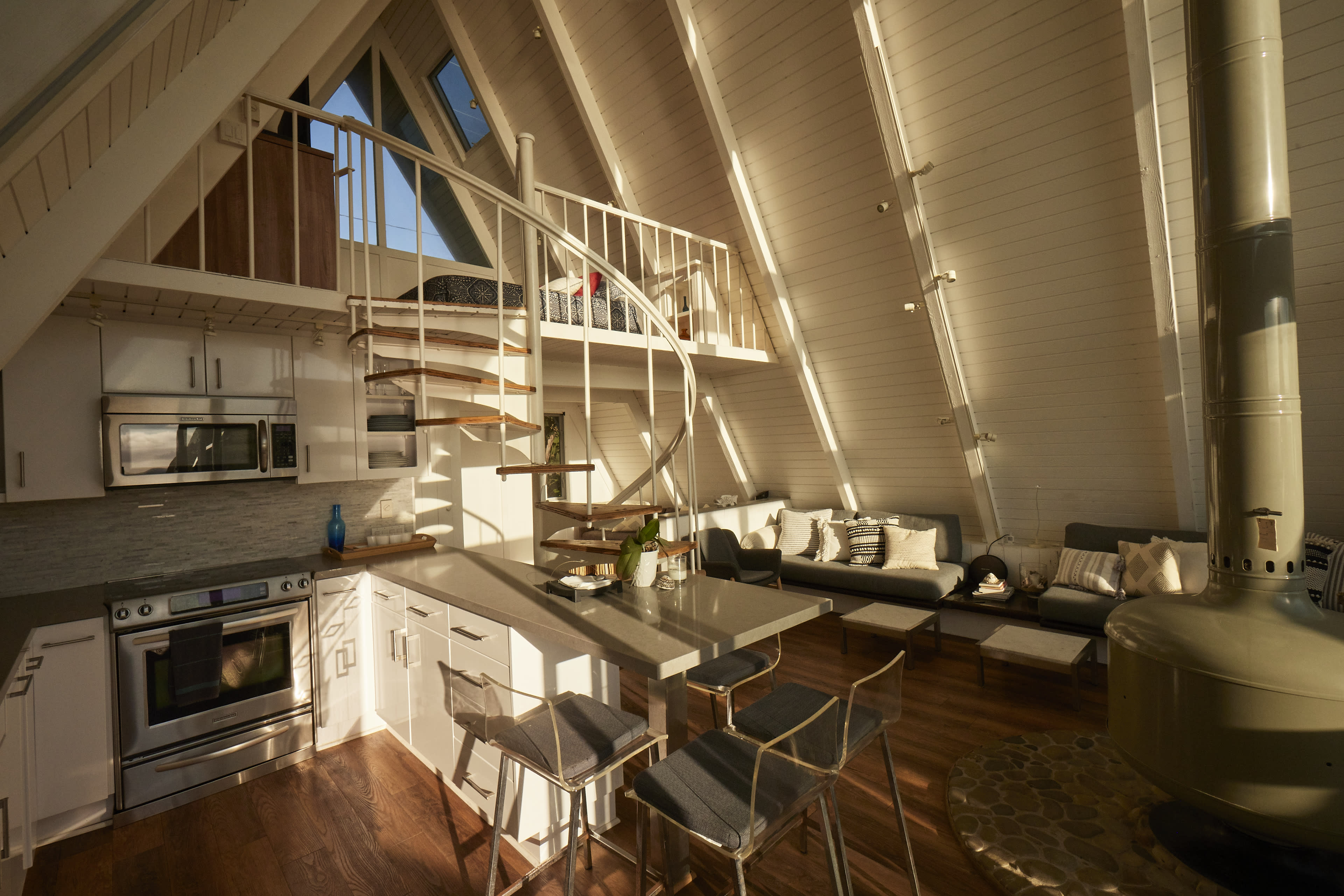 The image depicts a modern, A-frame interior with a spiral staircase, a kitchen island, and a seating area filled with light from large windows.