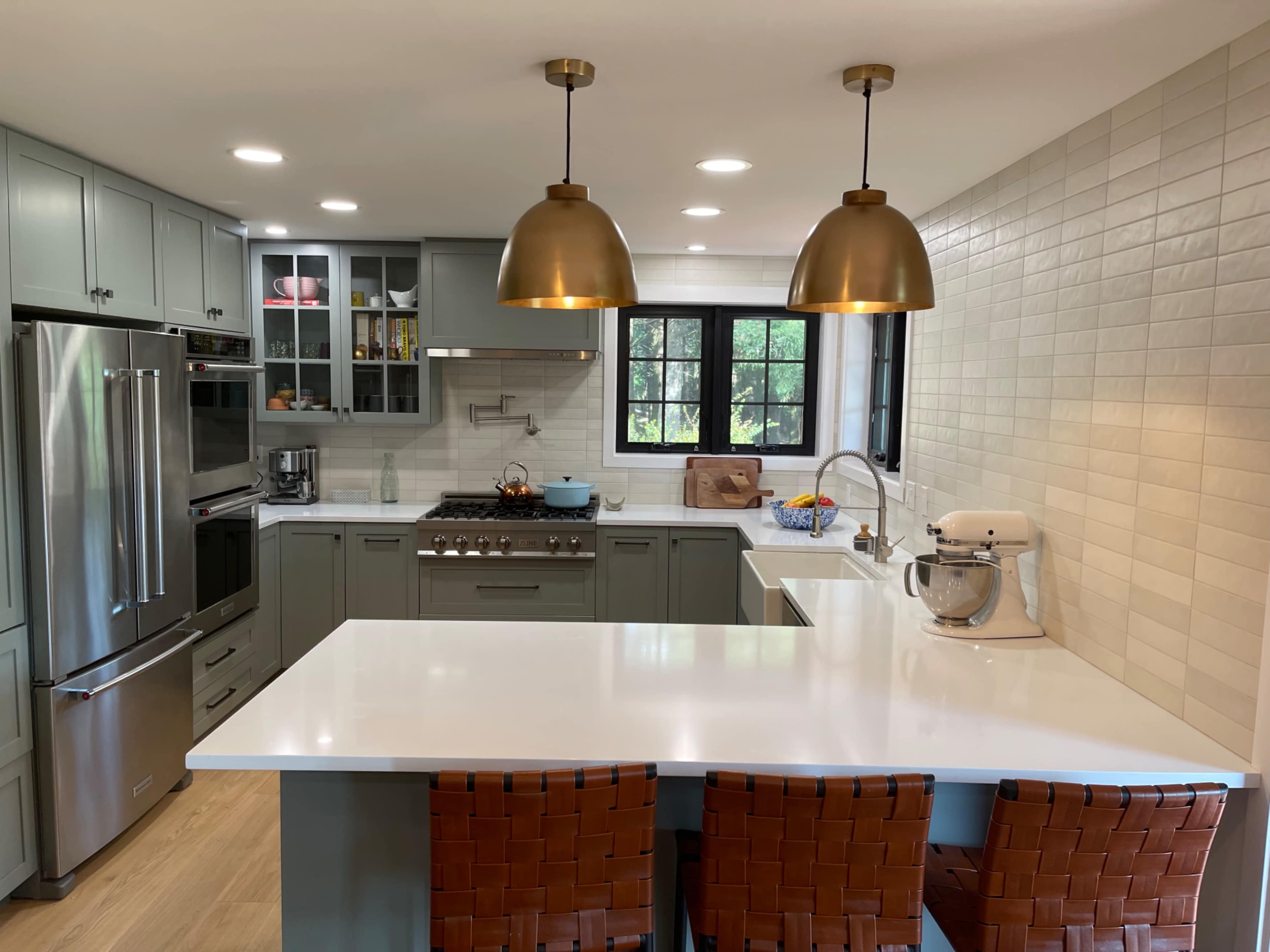 A modern kitchen features gray cabinetry, stainless steel appliances, and a white countertop with two pendant lights above a central island and three woven bar stools.