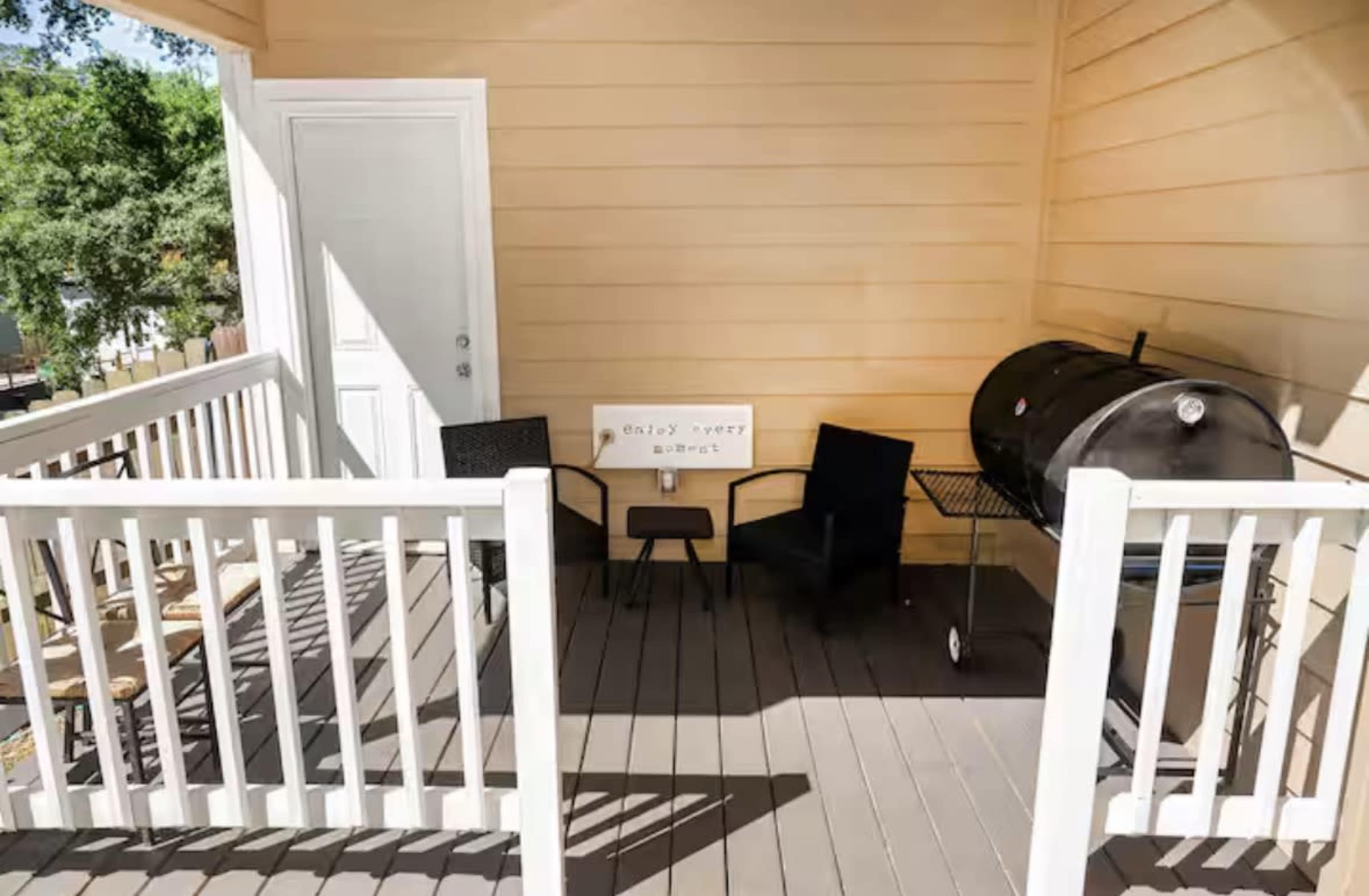 The image shows a small porch area with two black chairs, a sign, and a grill on a wooden deck.