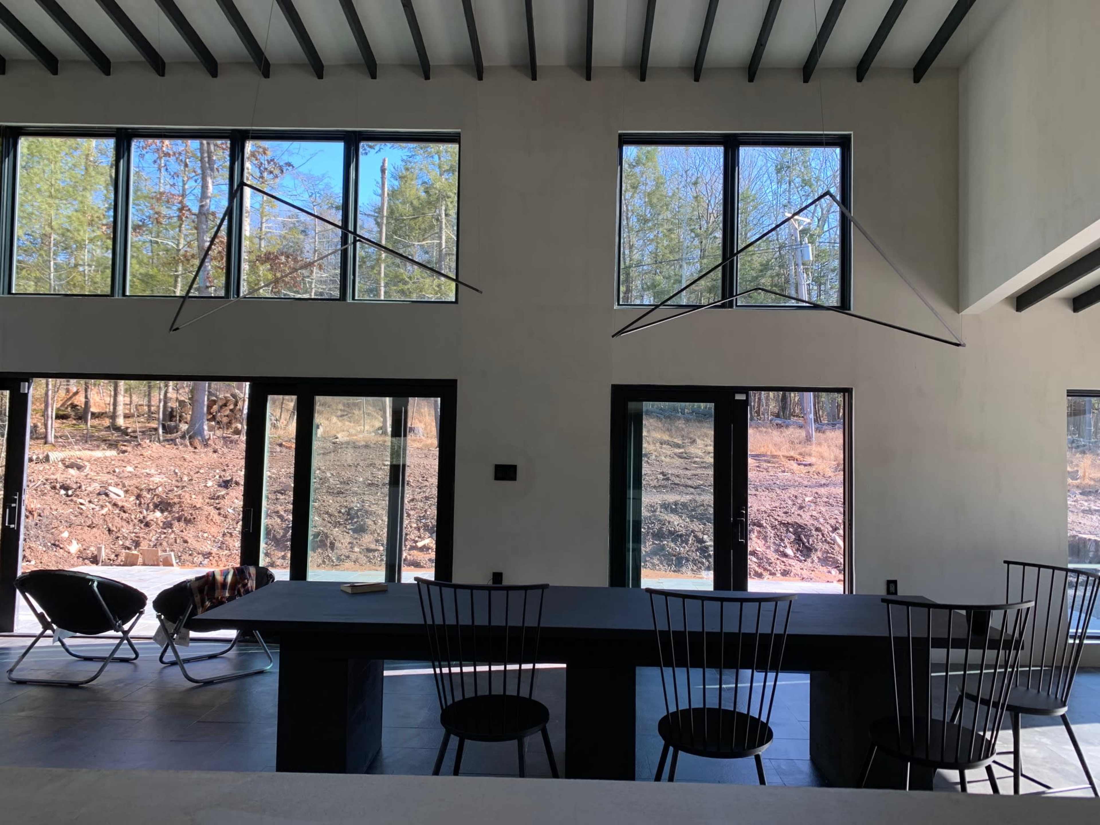 A modern, minimalistic interior featuring large windows overlooking a forested area, with a long black table and several wire-frame chairs arranged around it.