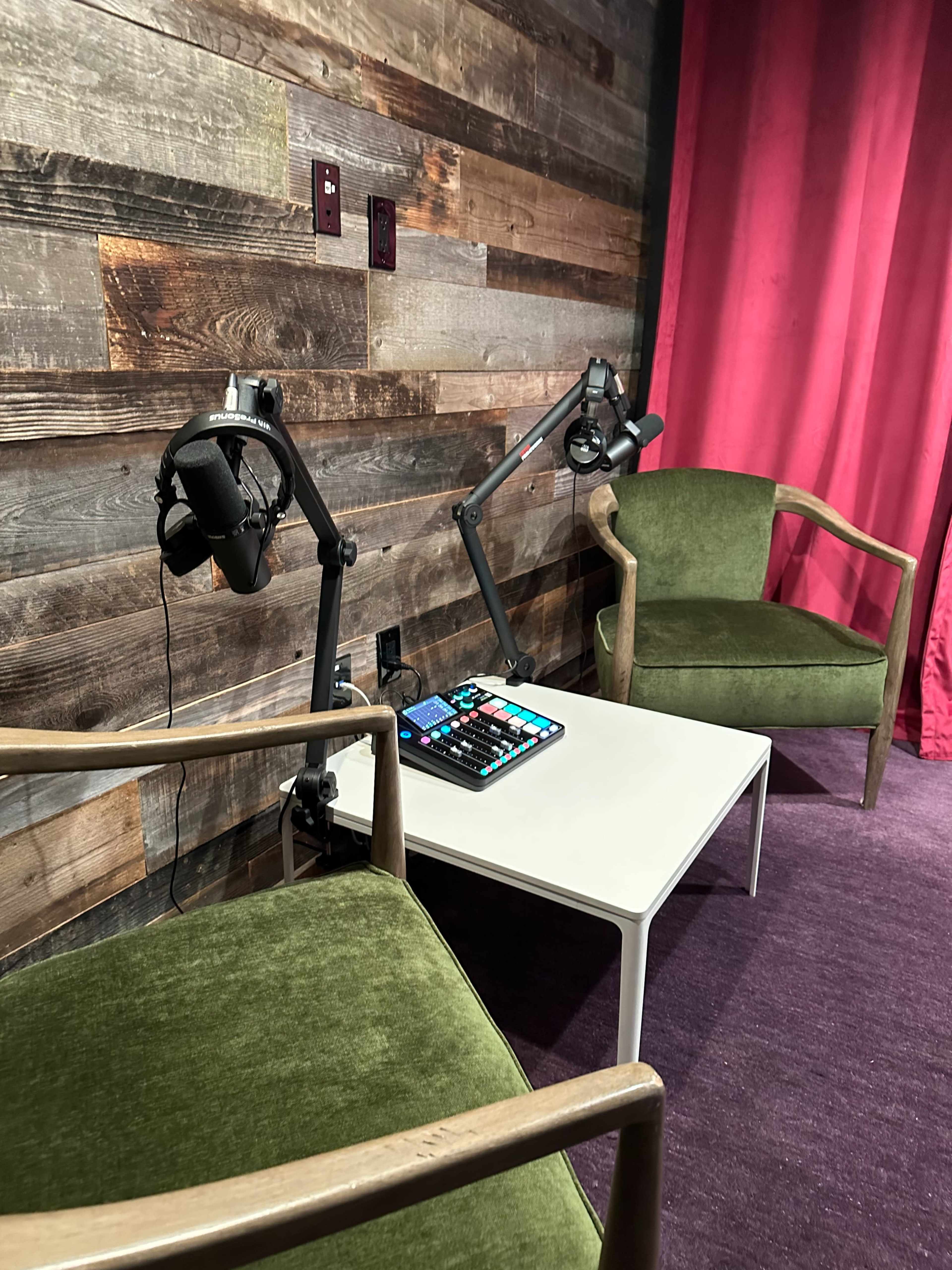 Two green chairs positioned around a white table in a room with wooden walls and pink curtains, with microphones mounted on adjustable arms and a control panel on the table.