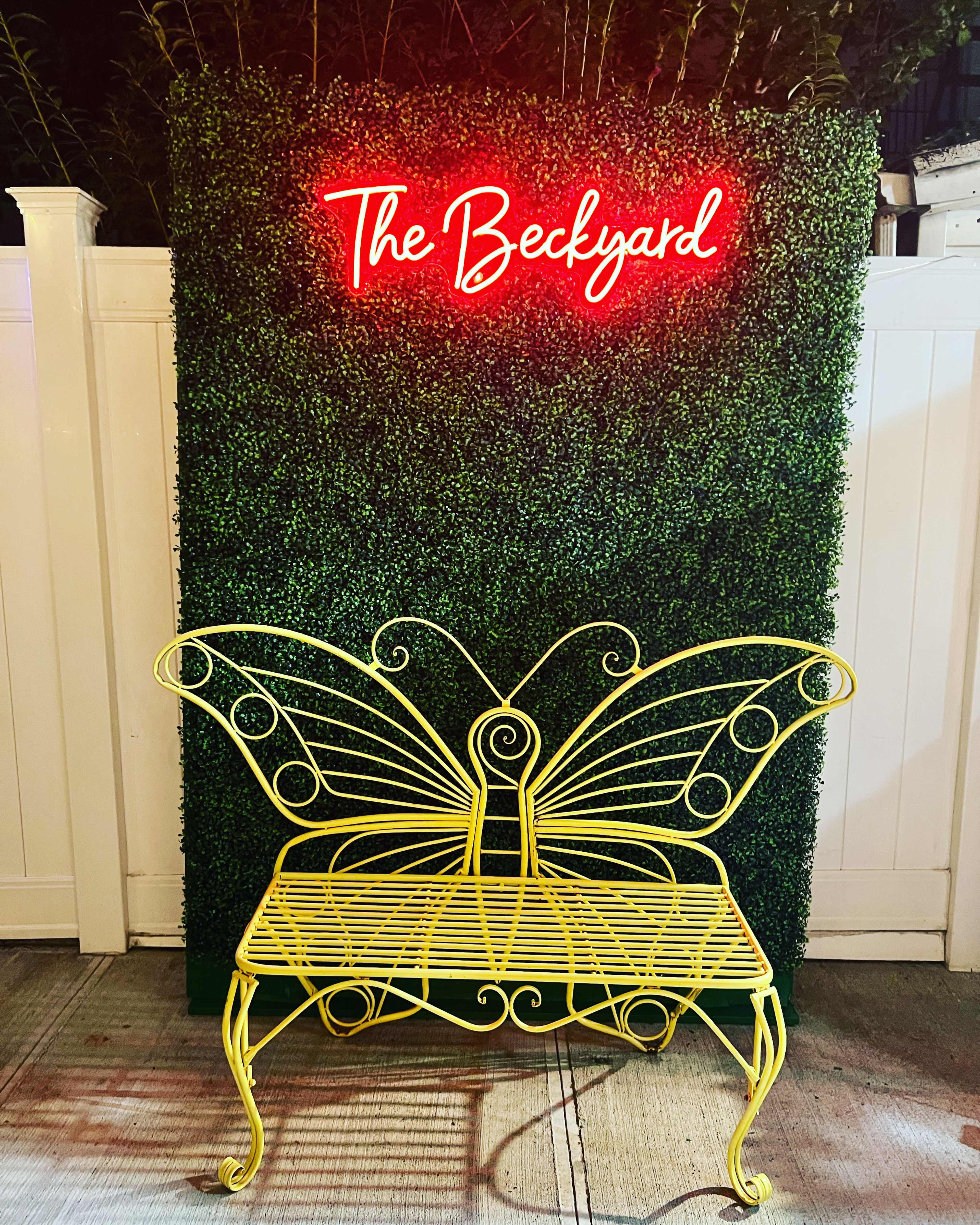 A yellow butterfly-shaped bench is positioned in front of a green hedge with a glowing red sign that reads "The Backyard."