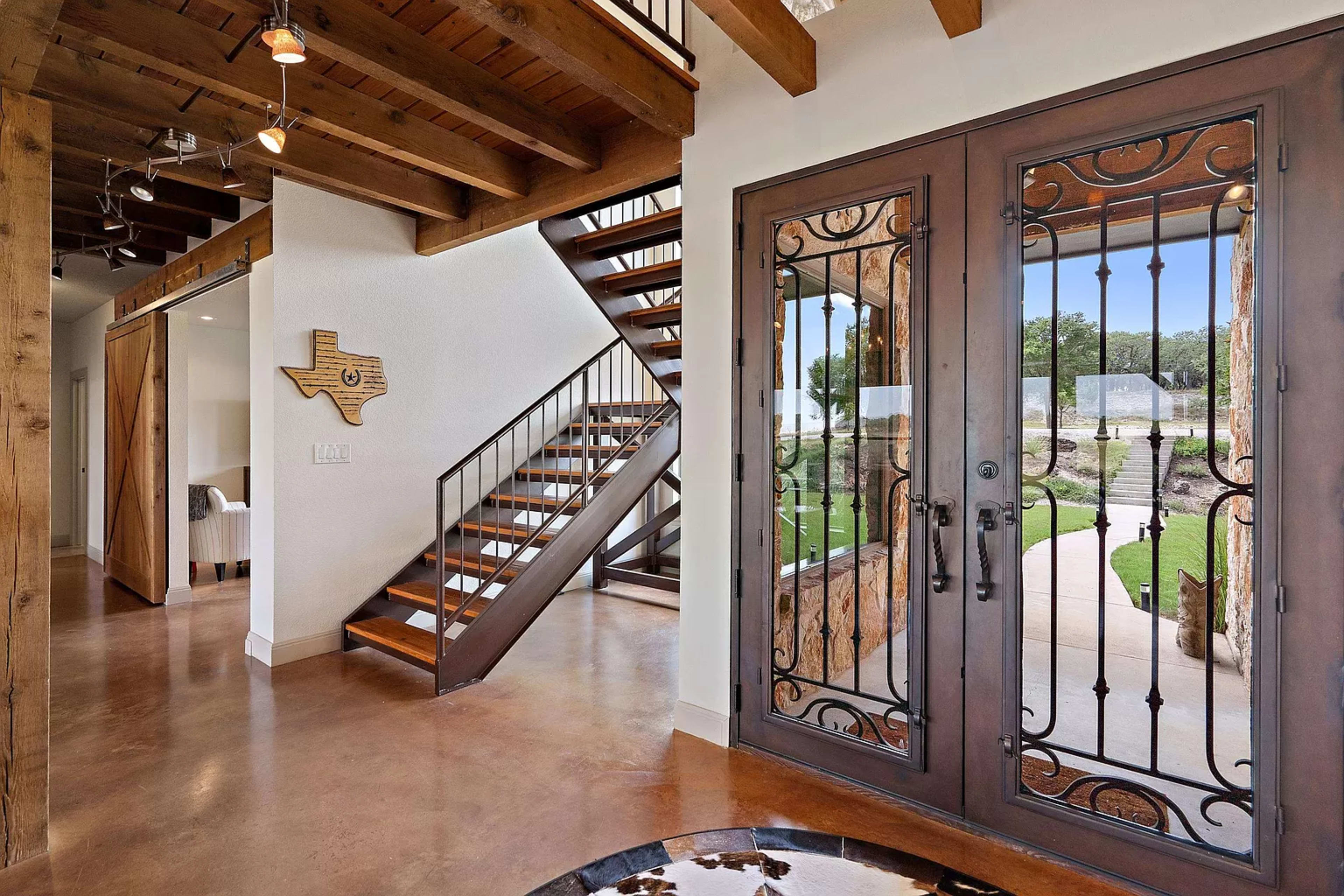 The image shows a spacious entryway with a wooden staircase, wrought iron doors, and a decorative Texas map on the wall.