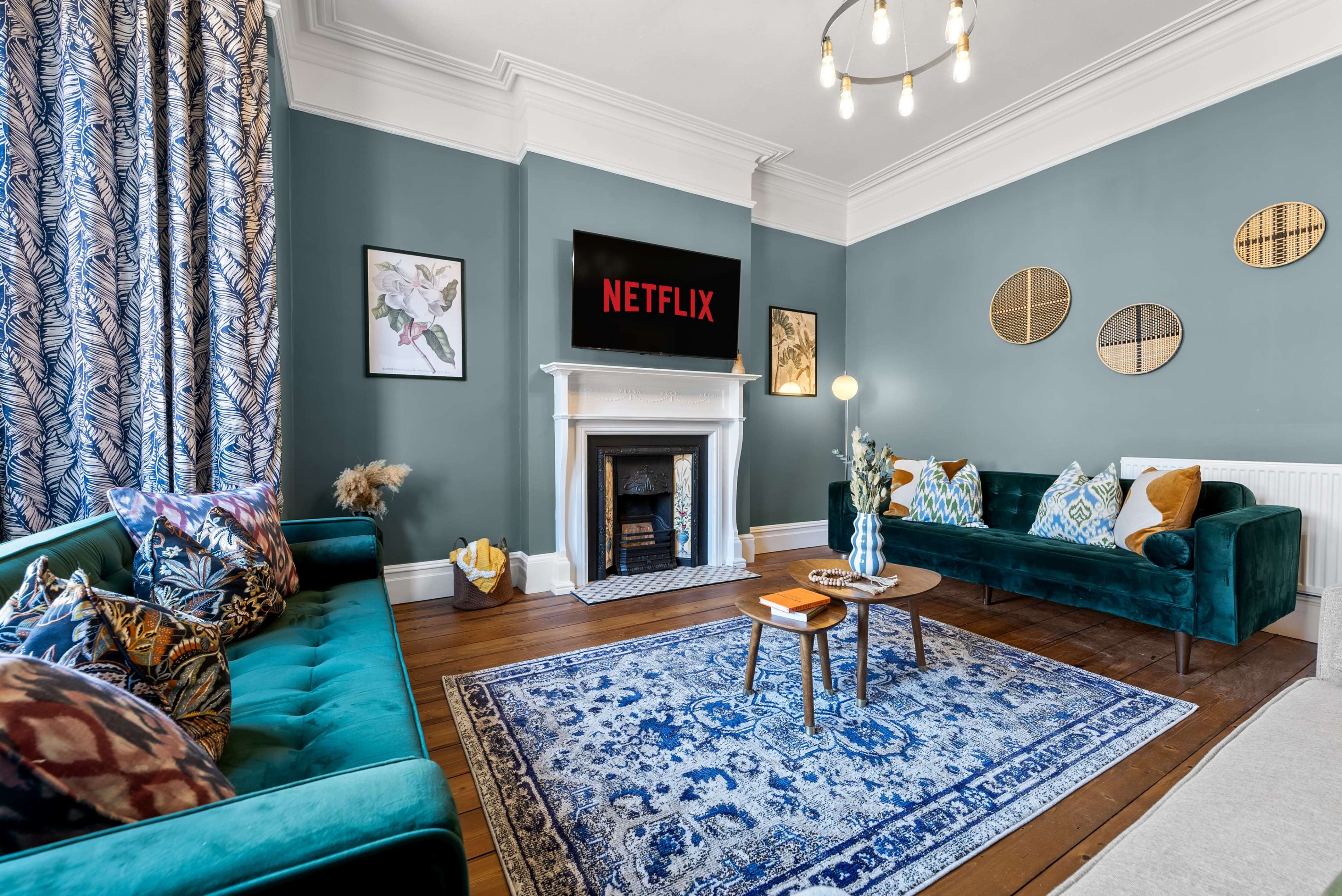 A cozy living room features two teal sofas, a coffee table, a decorative rug, and a wall-mounted television displaying the Netflix logo.
