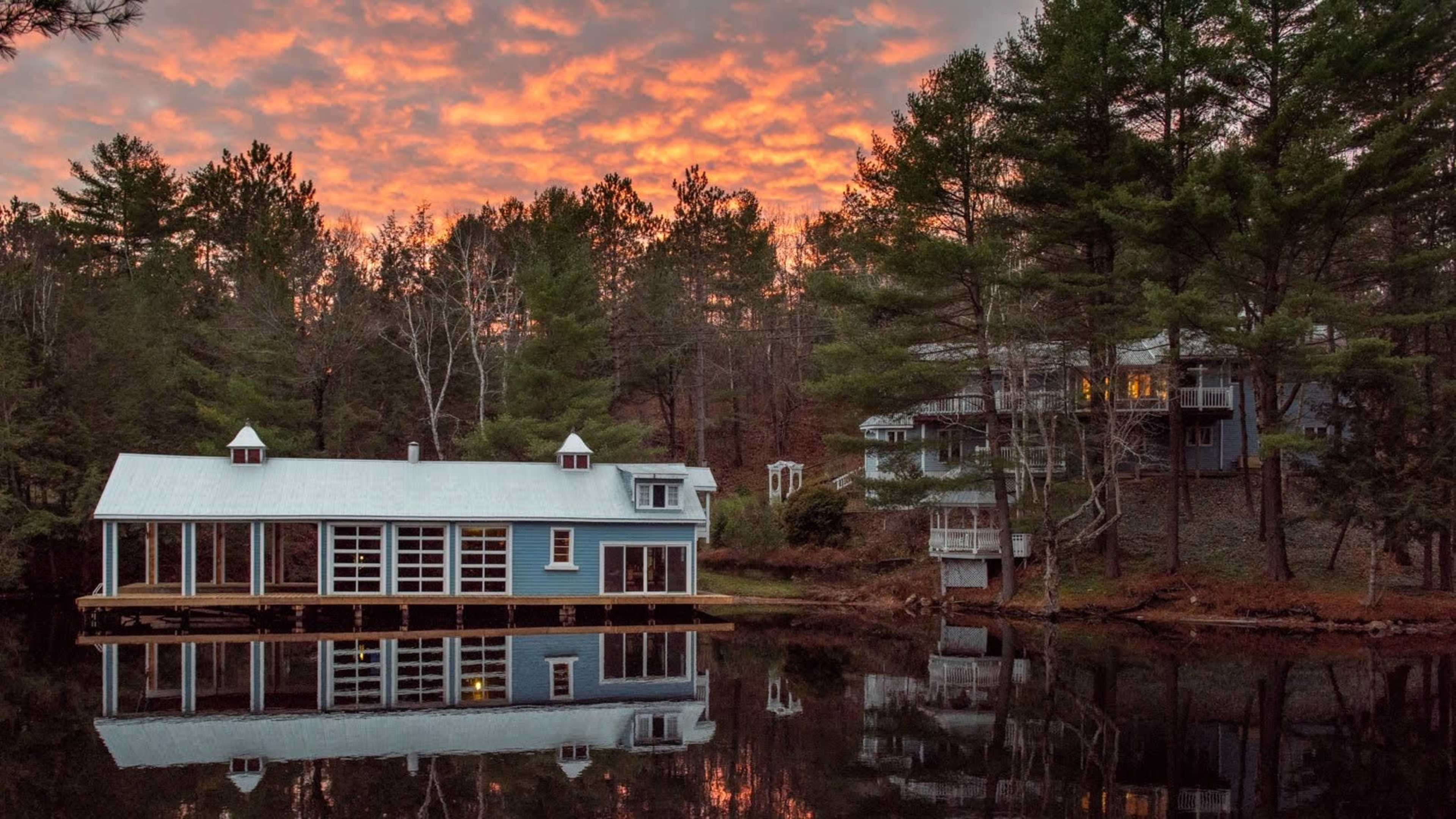 A blue house with a white roof sits next to a reflective pond, surrounded by trees and a bright orange sky at sunset.