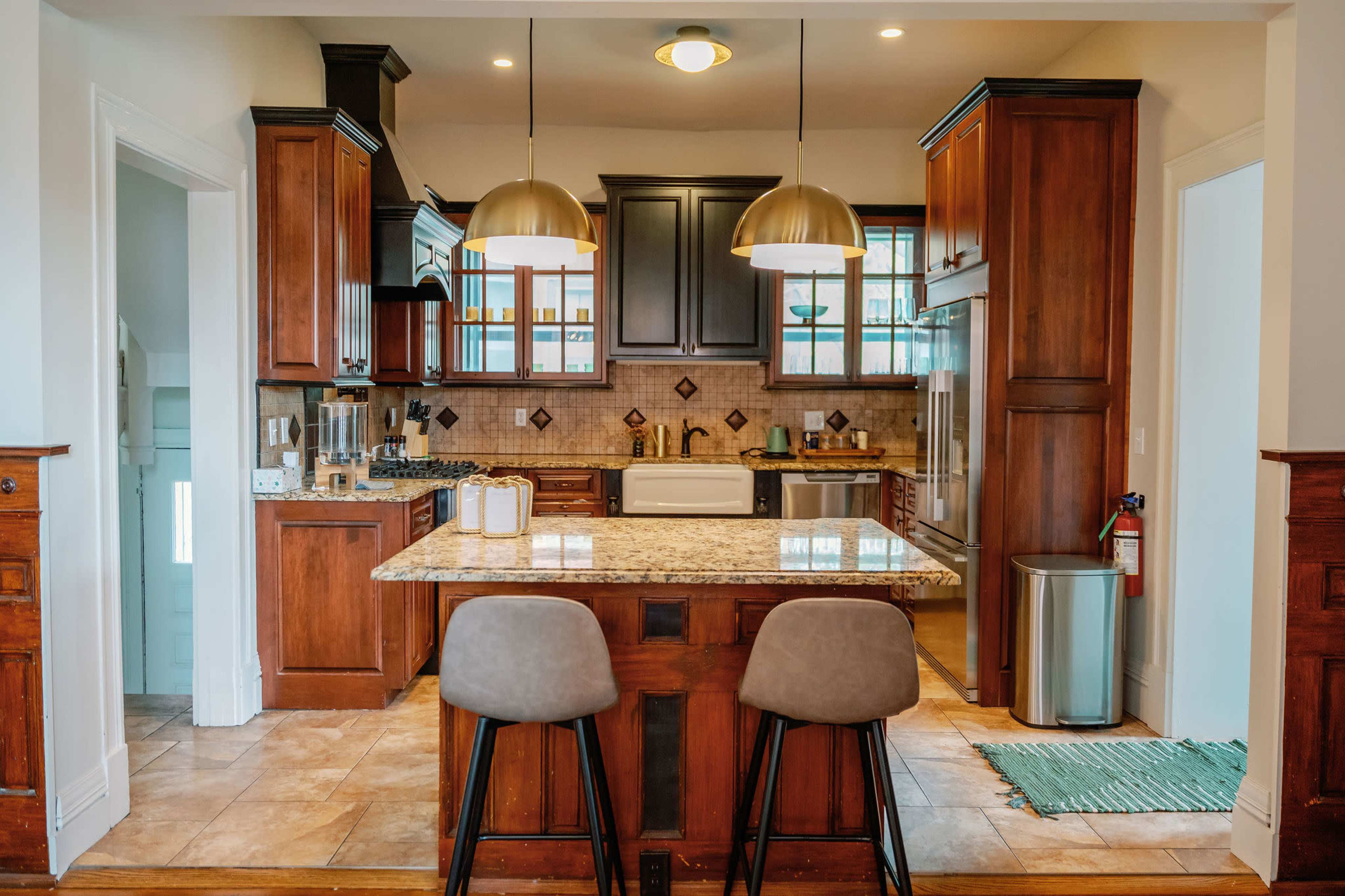 A modern kitchen features wooden cabinets, a large island with two stools, and pendant lighting above.