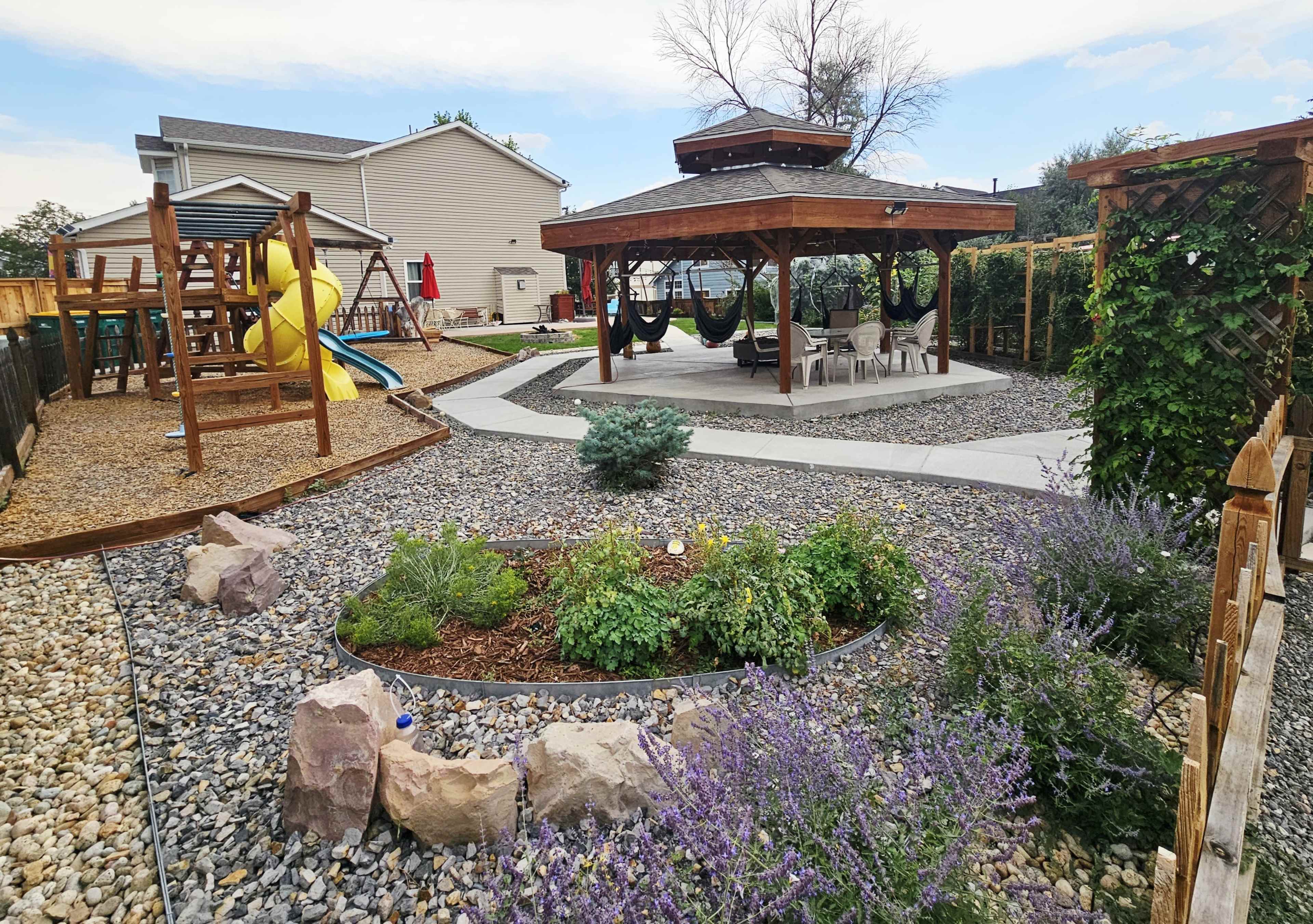 The image shows a landscaped backyard featuring a gazebo with seating, a playground equipment area, and various plants and stones.