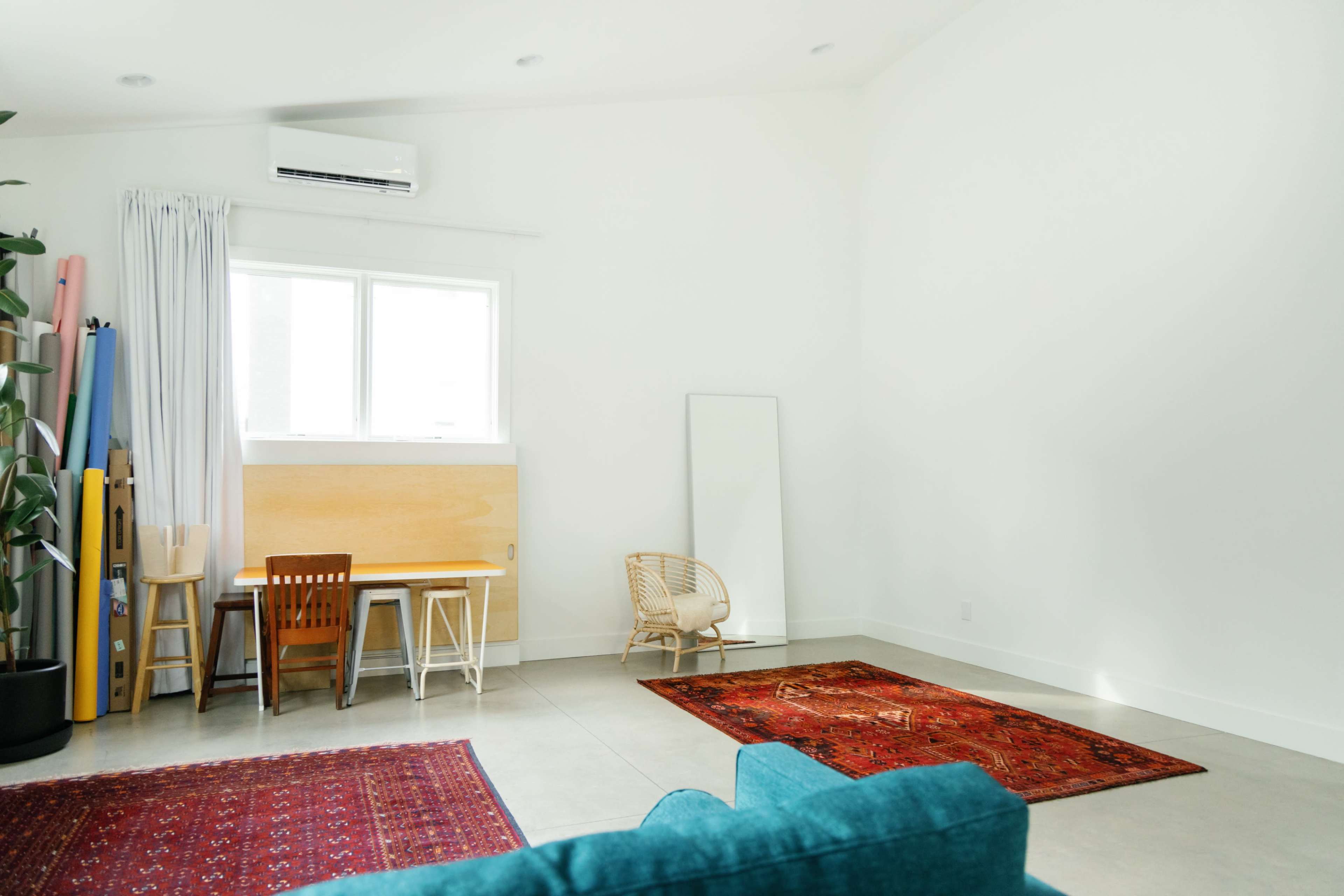 The image shows a minimalist indoor space featuring a light-colored wall, a dining area with a table and chairs, a large mirror, and a few rugs on the floor.