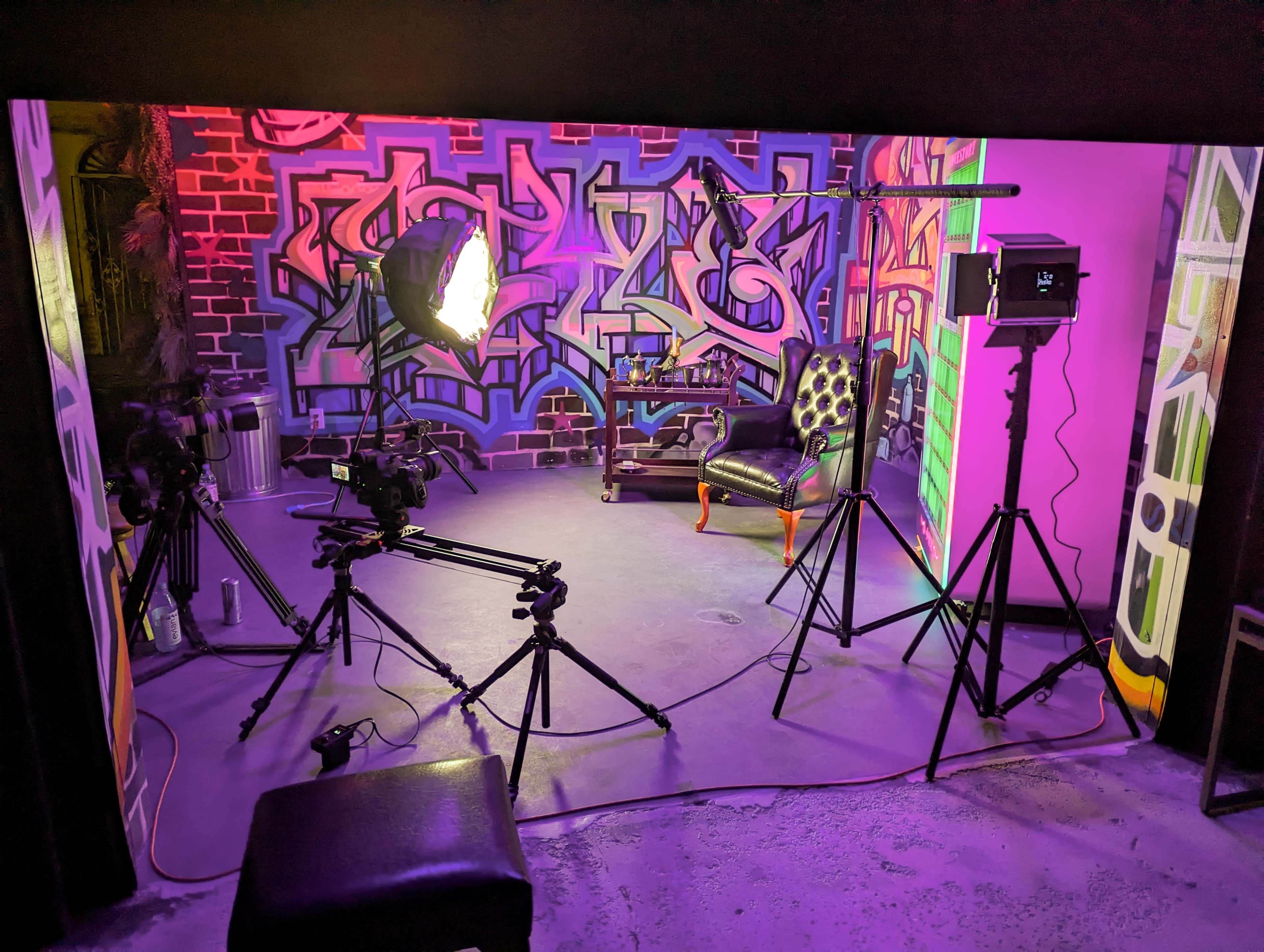 A photography studio setup with various lights and cameras positioned around a black chair in front of a colorful graffiti backdrop.