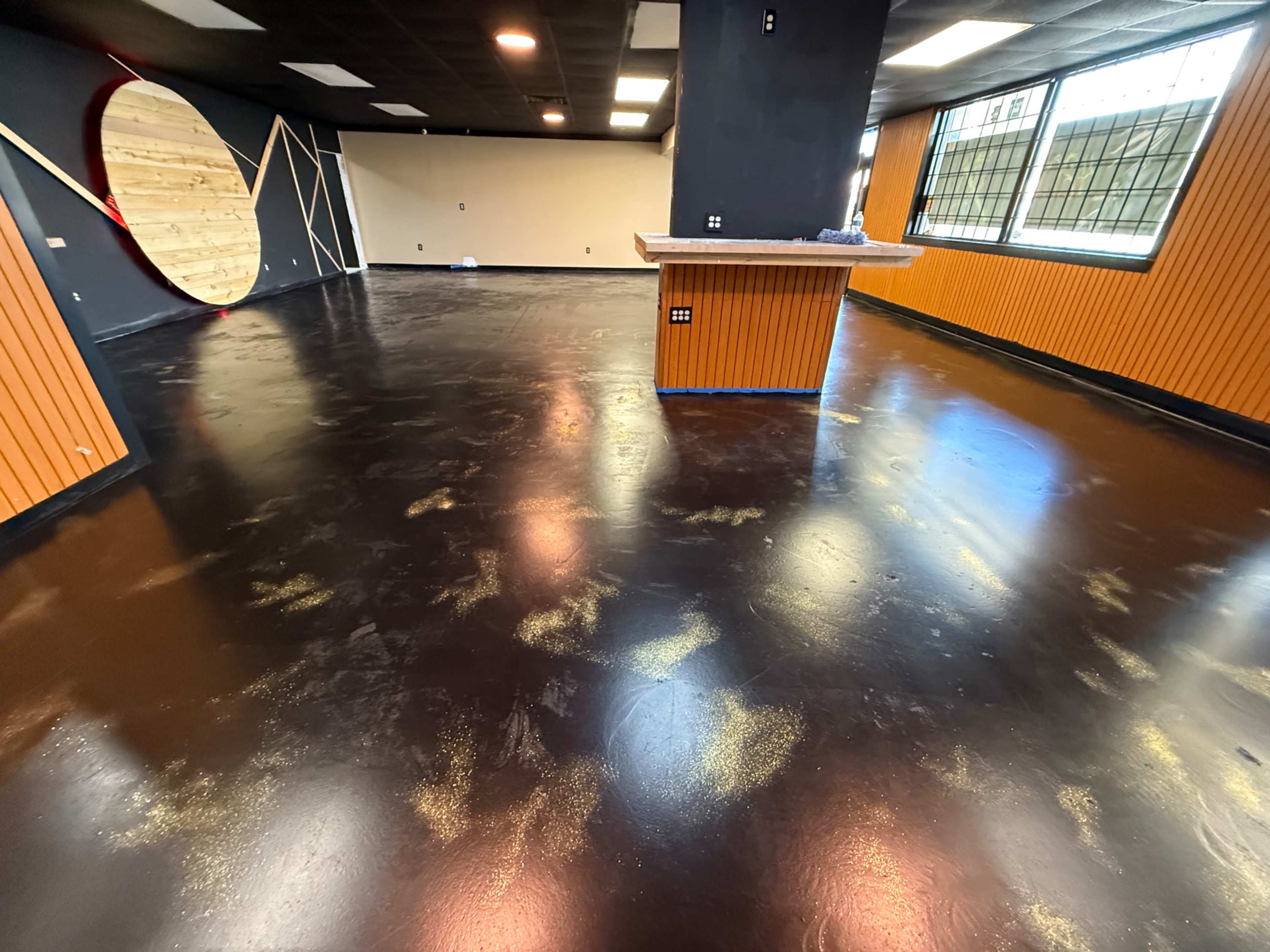 The image shows a spacious interior room with a glossy black floor, wooden paneling on the walls, and a partially finished bar area.