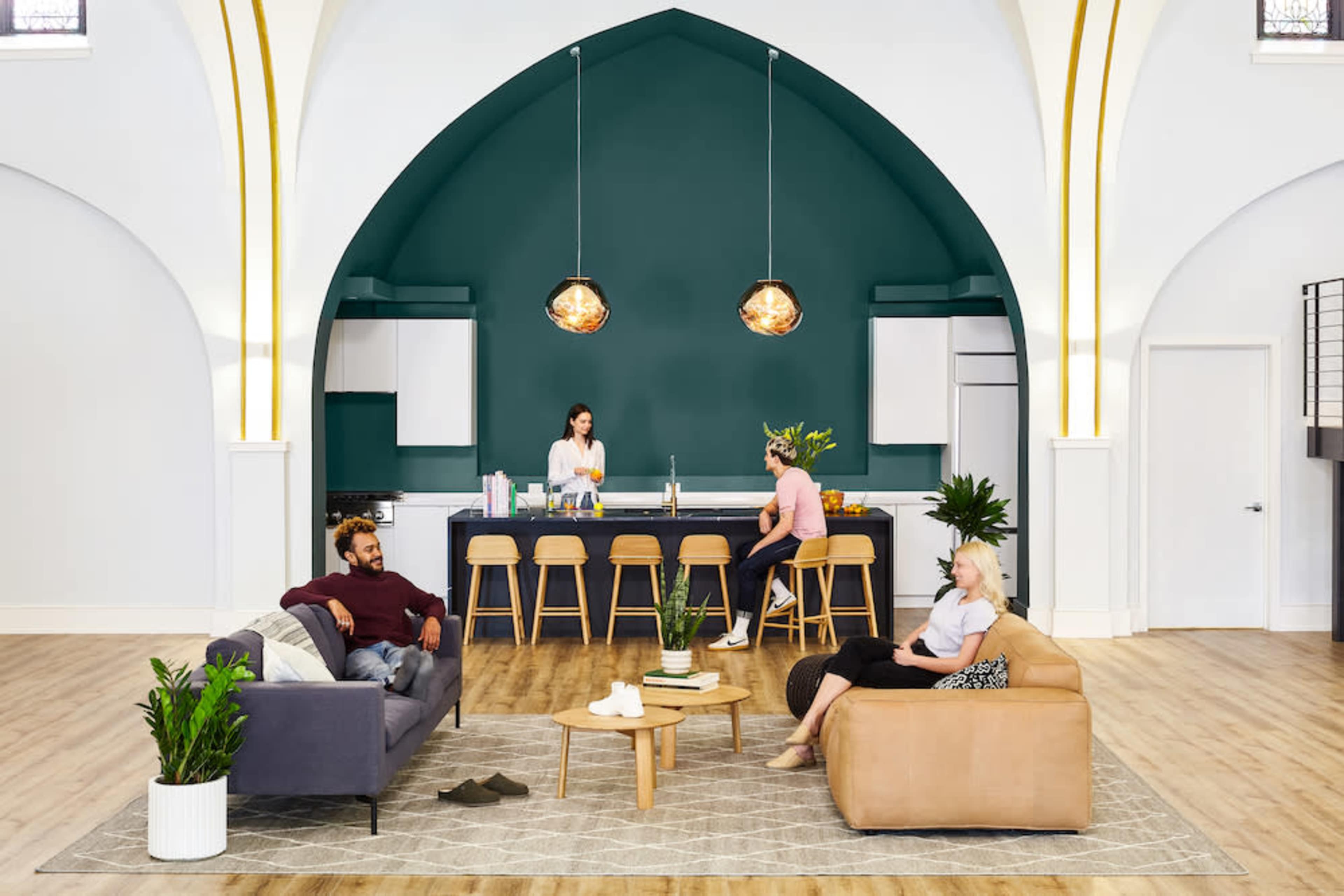 A bright and spacious living area features a kitchen with bar stools, while two people relax on a sofa and an armchair, and a third person prepares food at the counter.