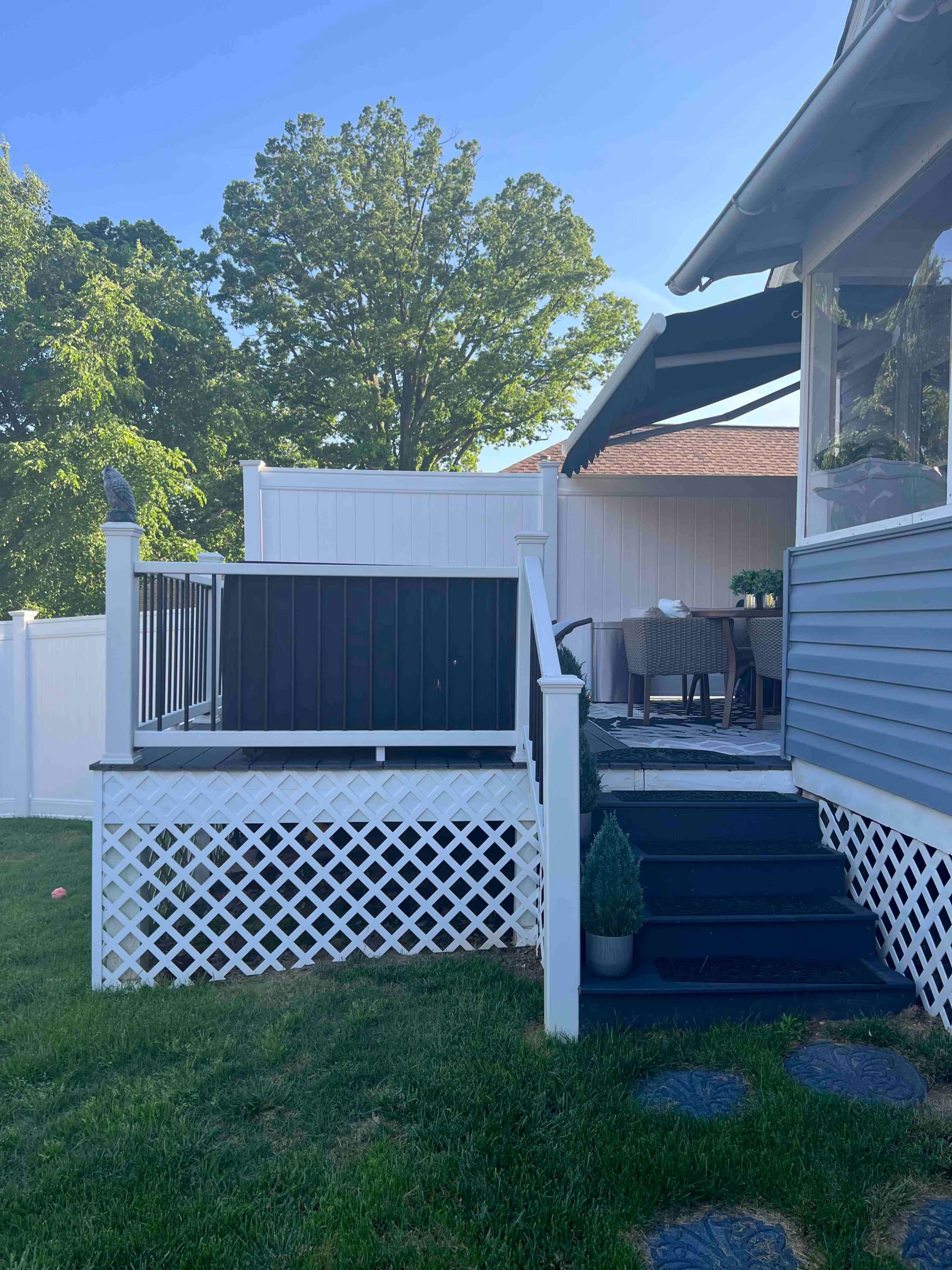 The image shows a backyard deck with a black railing and a table set for outdoor dining next to a white fence and green grass.