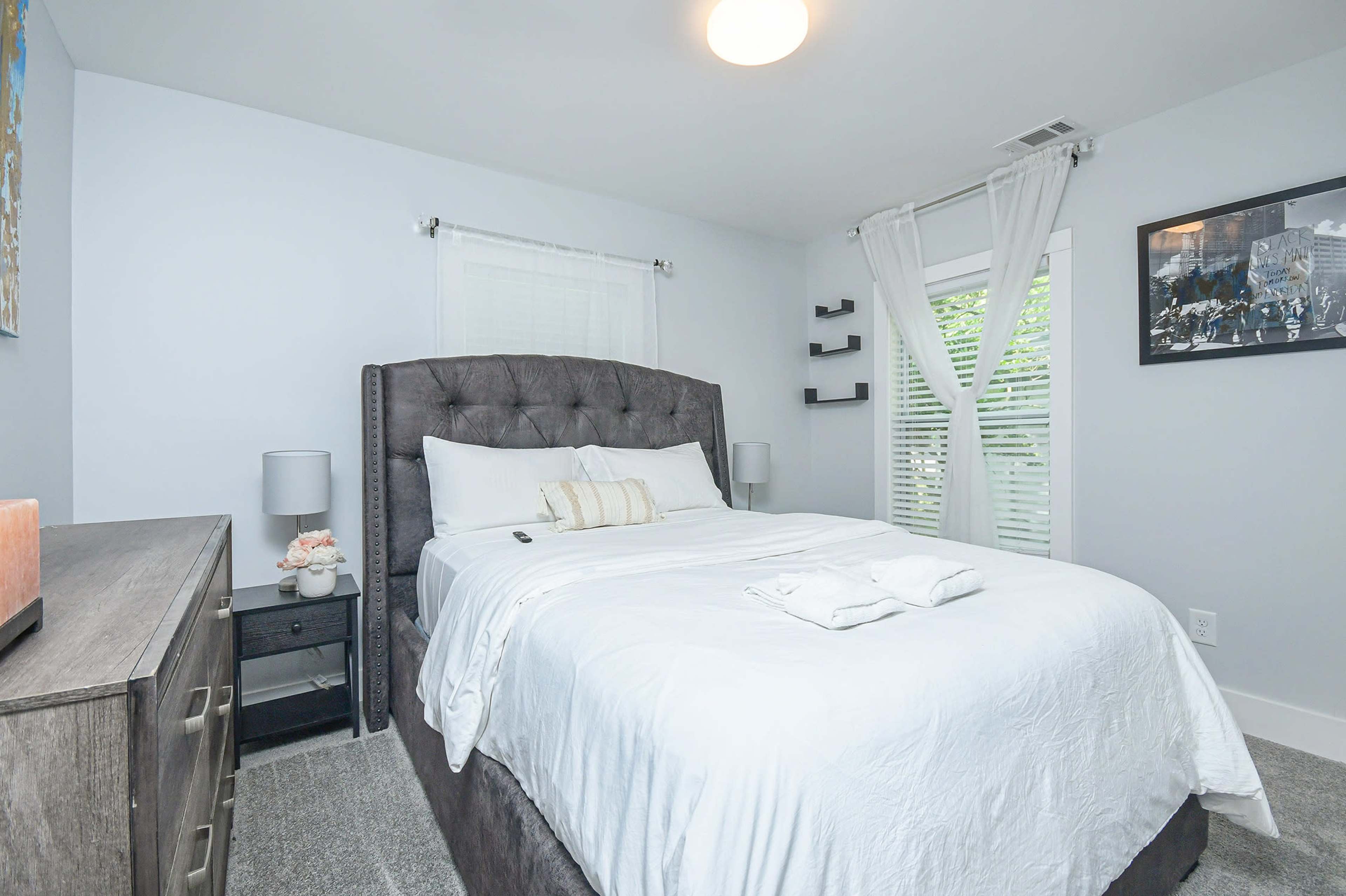 A cozy bedroom featuring a bed with a gray tufted headboard, light-colored bedding, a nightstand with lamps, and a dresser.