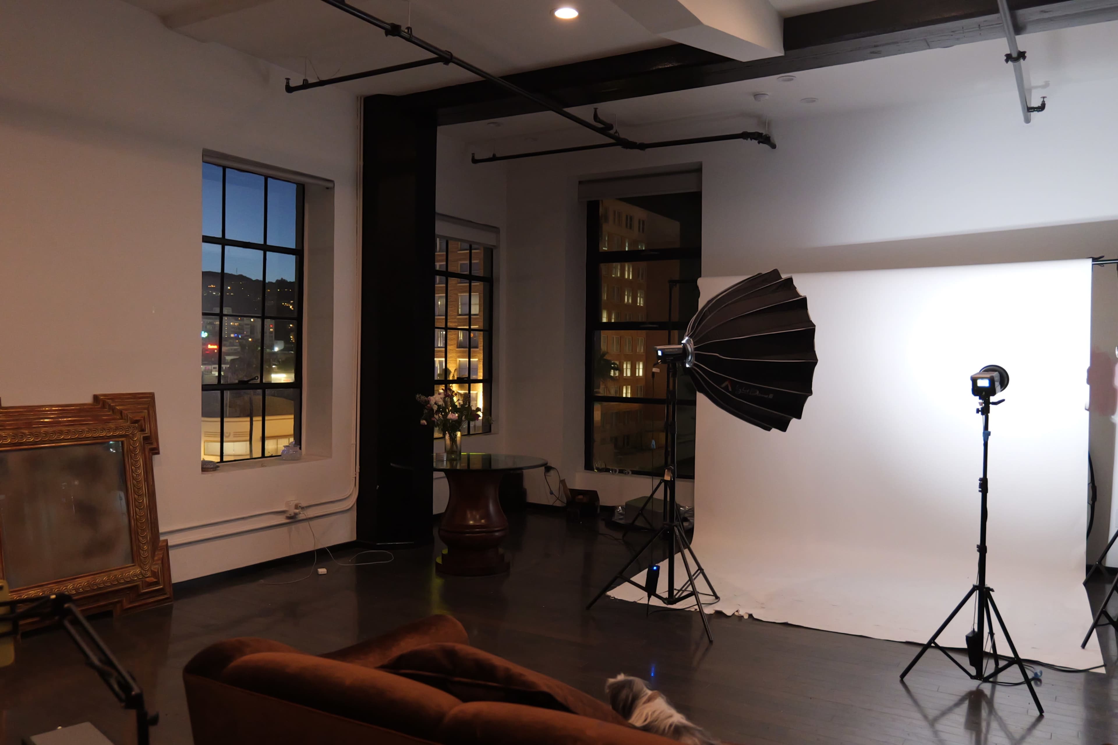 The image shows an interior space with a large window, a brown couch, a photography lighting setup, and a reflective mirror leaning against the wall.