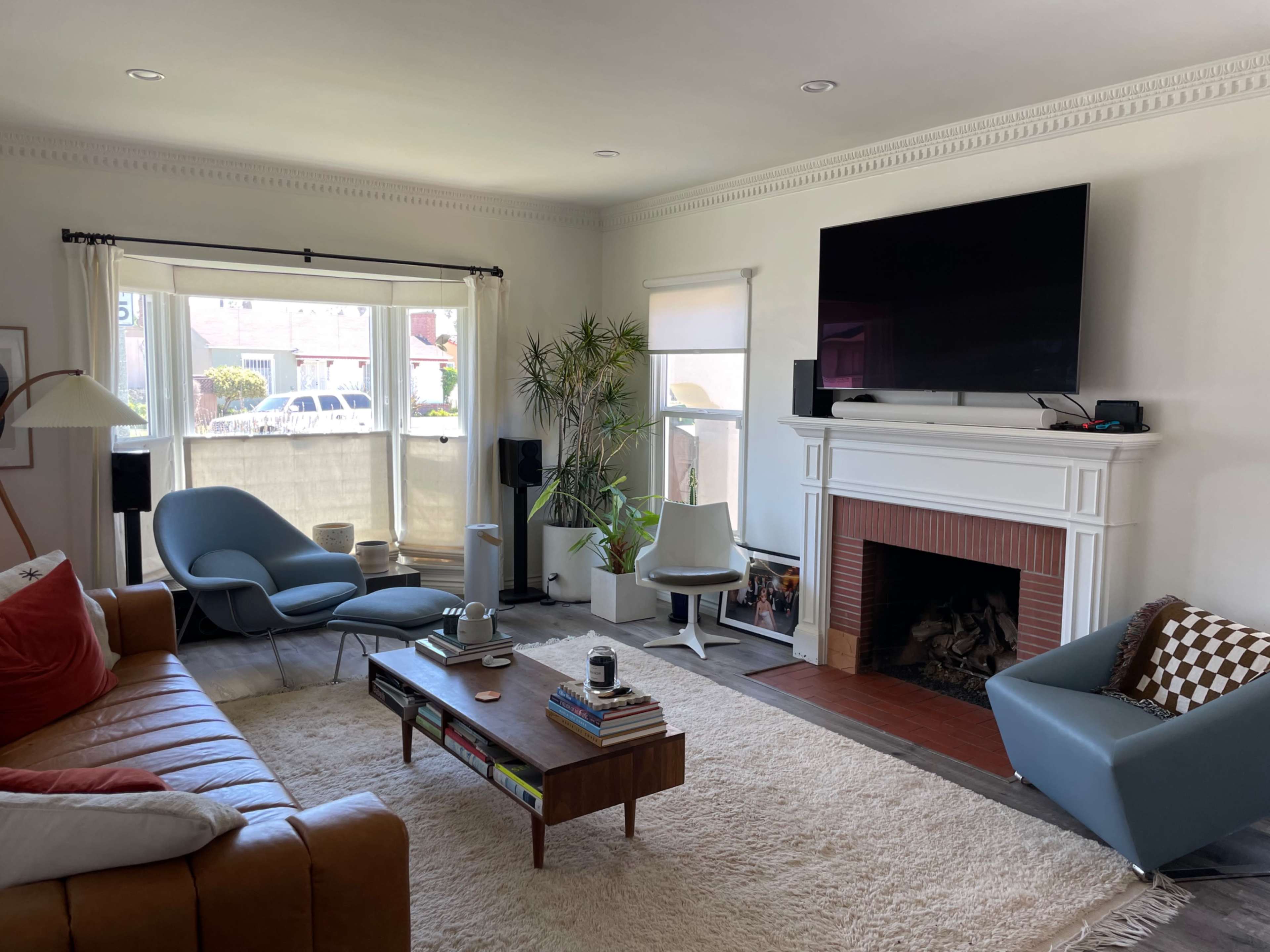 A furnished living room with a brown leather sofa, a coffee table, and a large television mounted above a white fireplace, all set against light-colored walls and large windows.