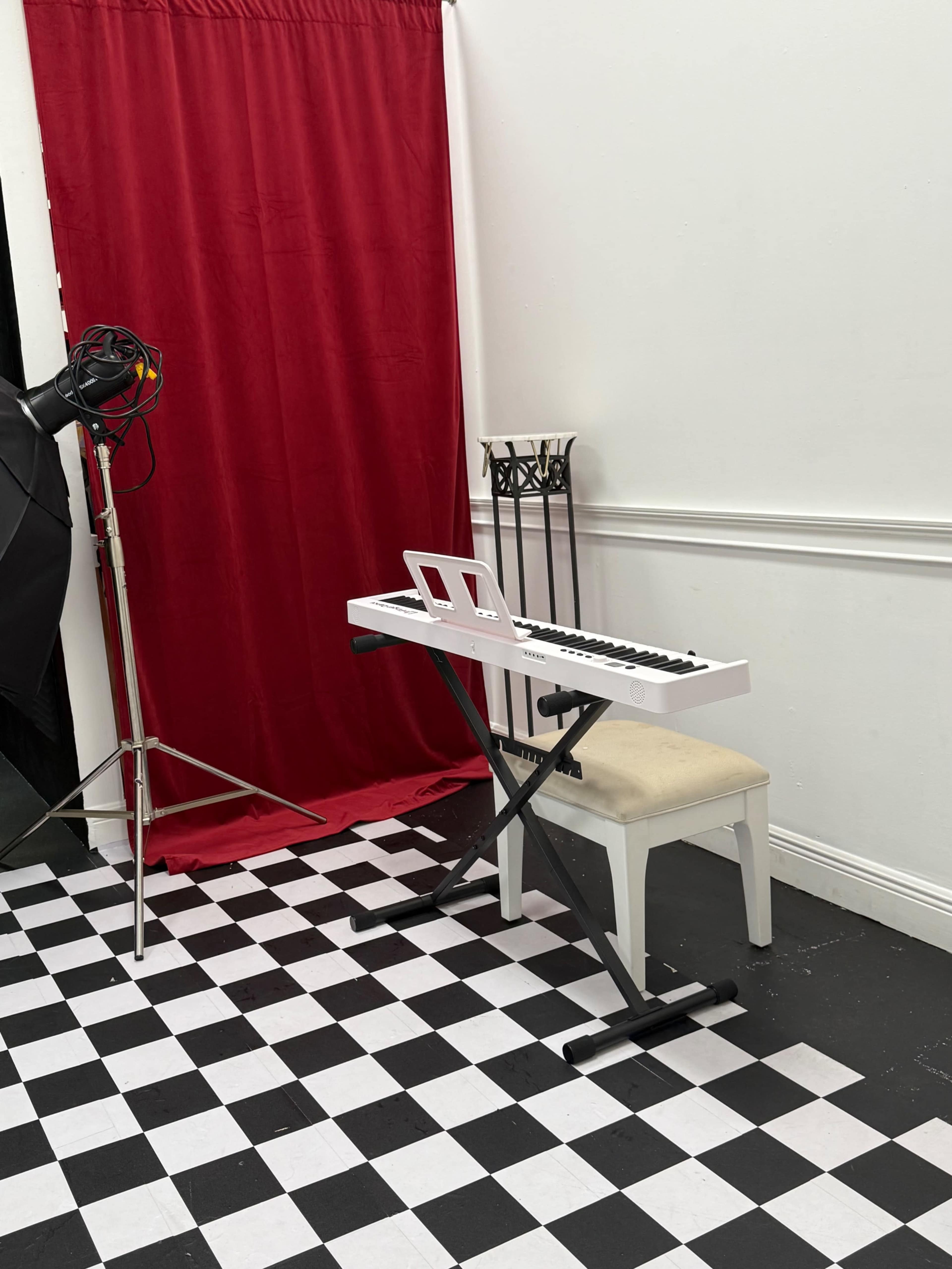 A white keyboard is placed on a black and white checkered floor in front of a red curtain, next to a tall chair and a lighting stand.
