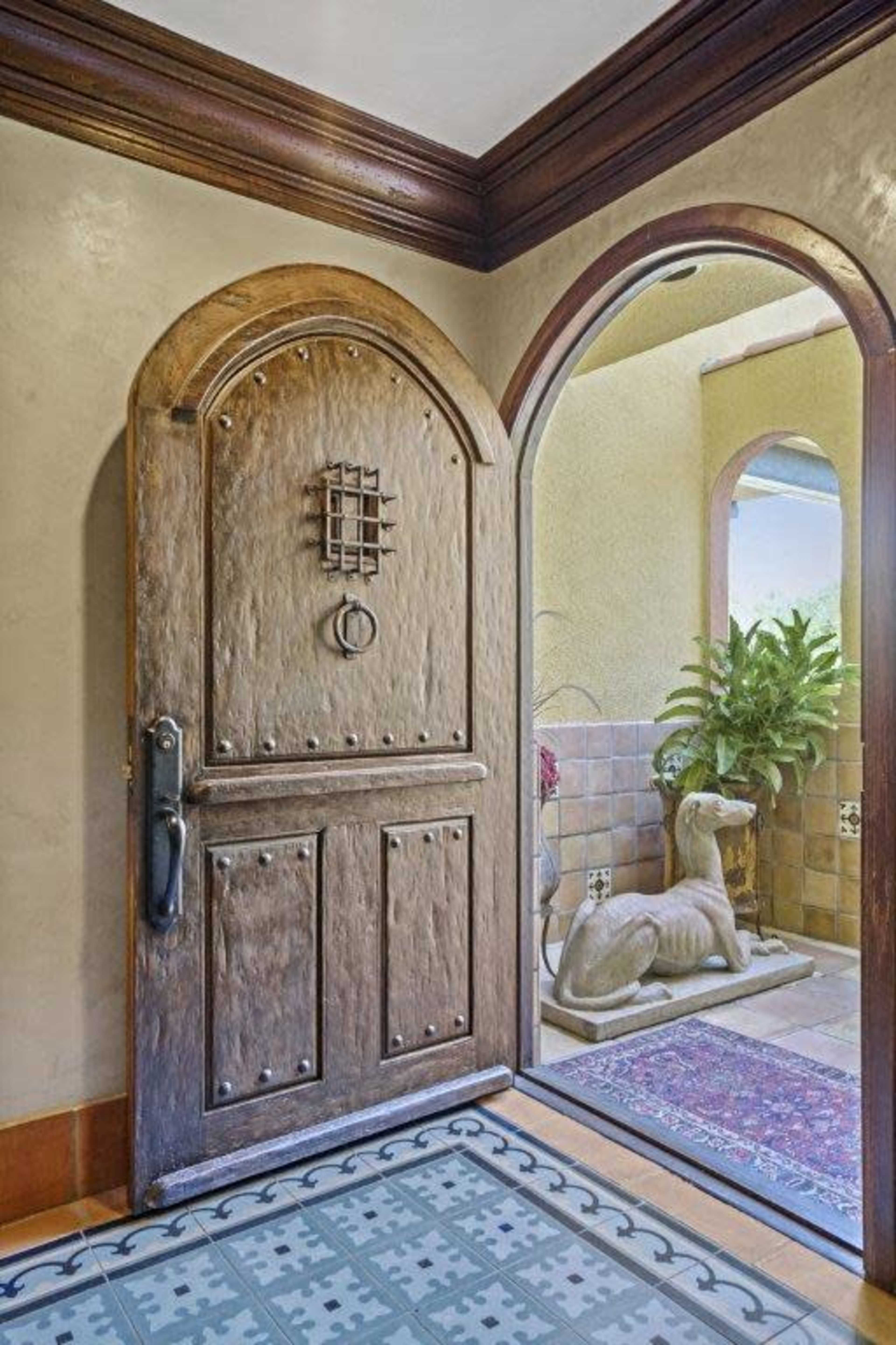 The image shows a wooden front door with a wrought iron grid and a circular handle, leading to an interior space featuring a statue of a dog and a decorative rug.