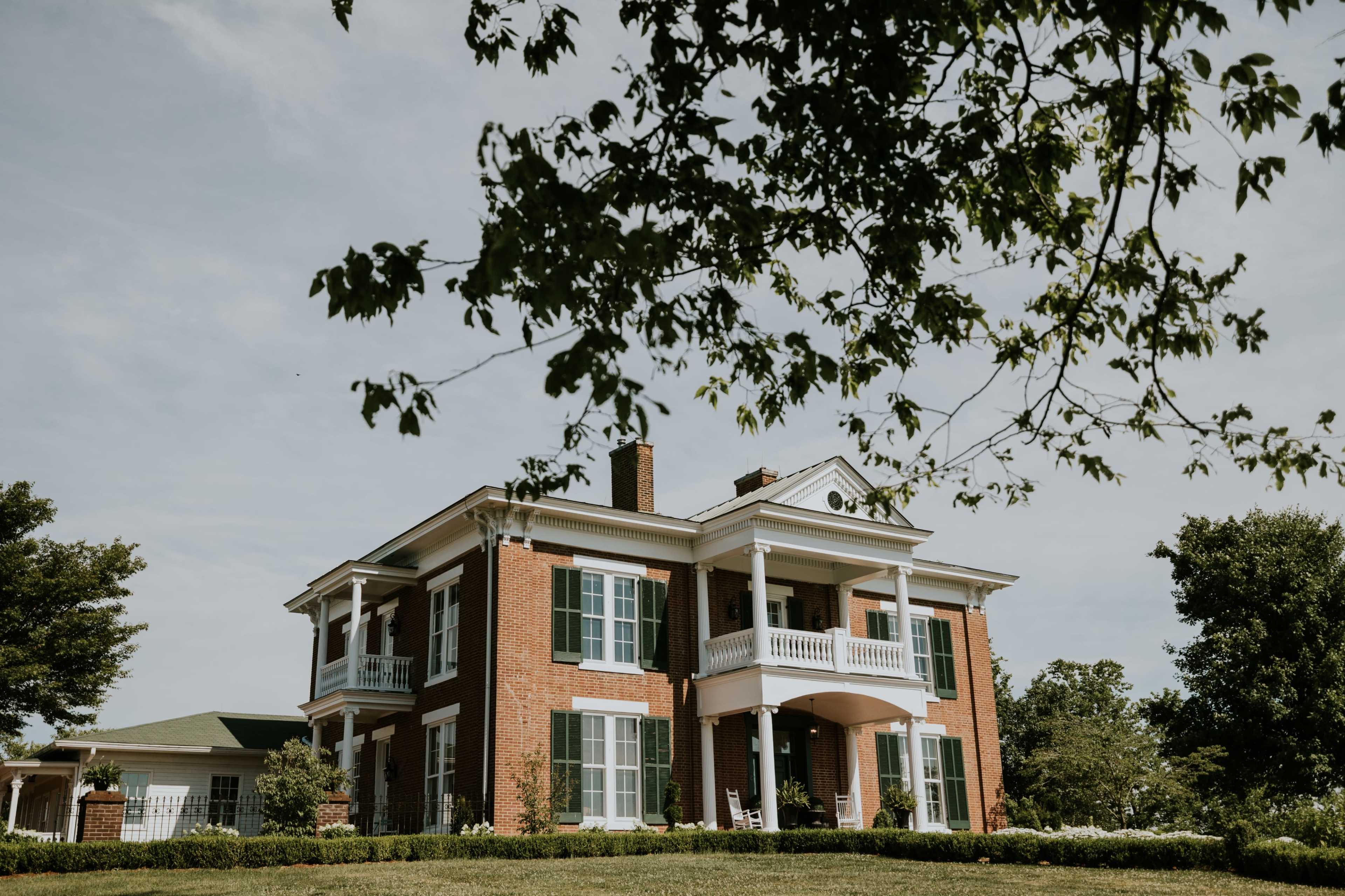 The image shows a large brick house with white trim and green shutters, surrounded by grass and trees.
