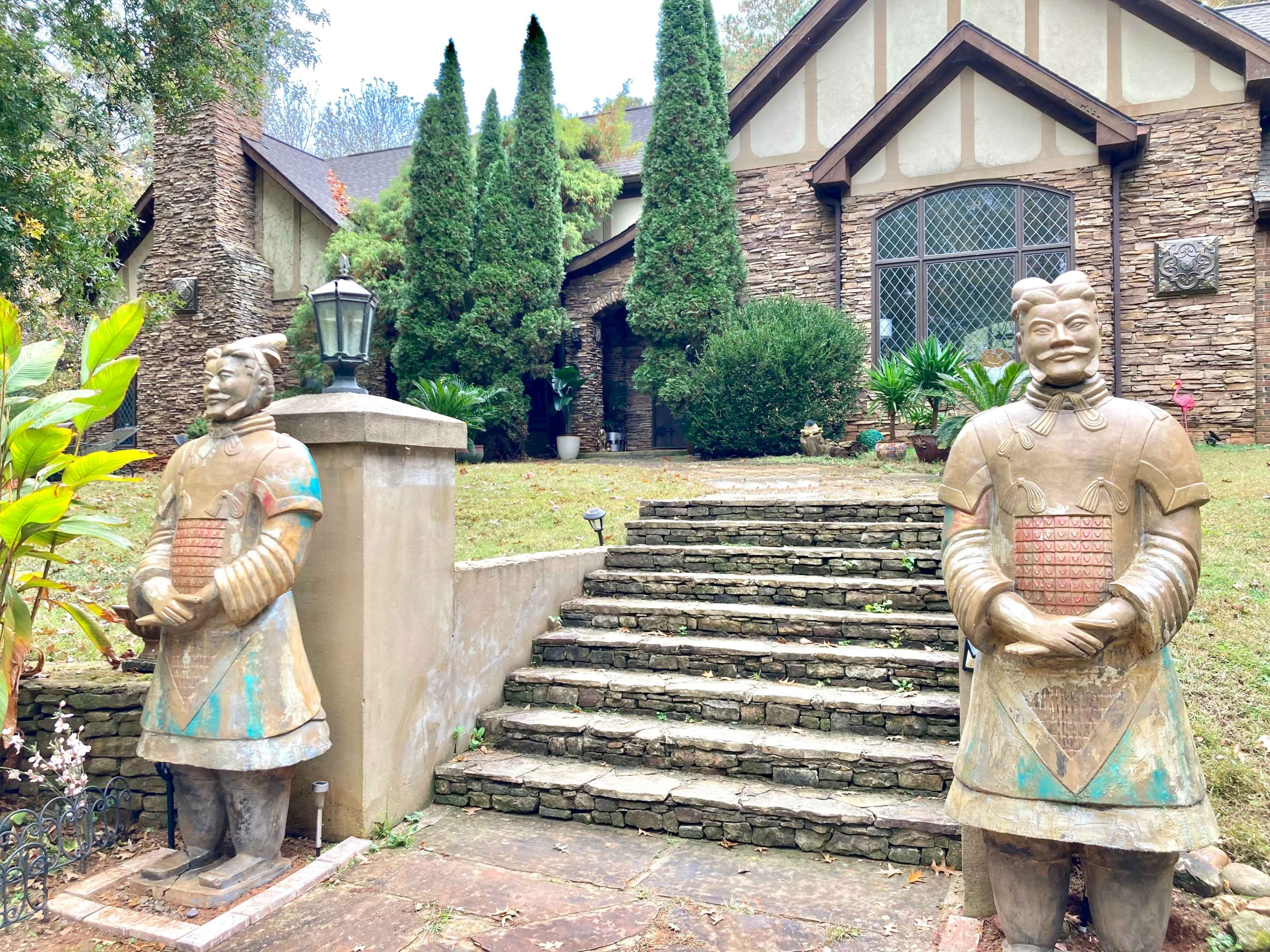 Two large stone warrior statues flank the entrance of a stone-clad house with a landscaped pathway and tall, narrow trees in the background.