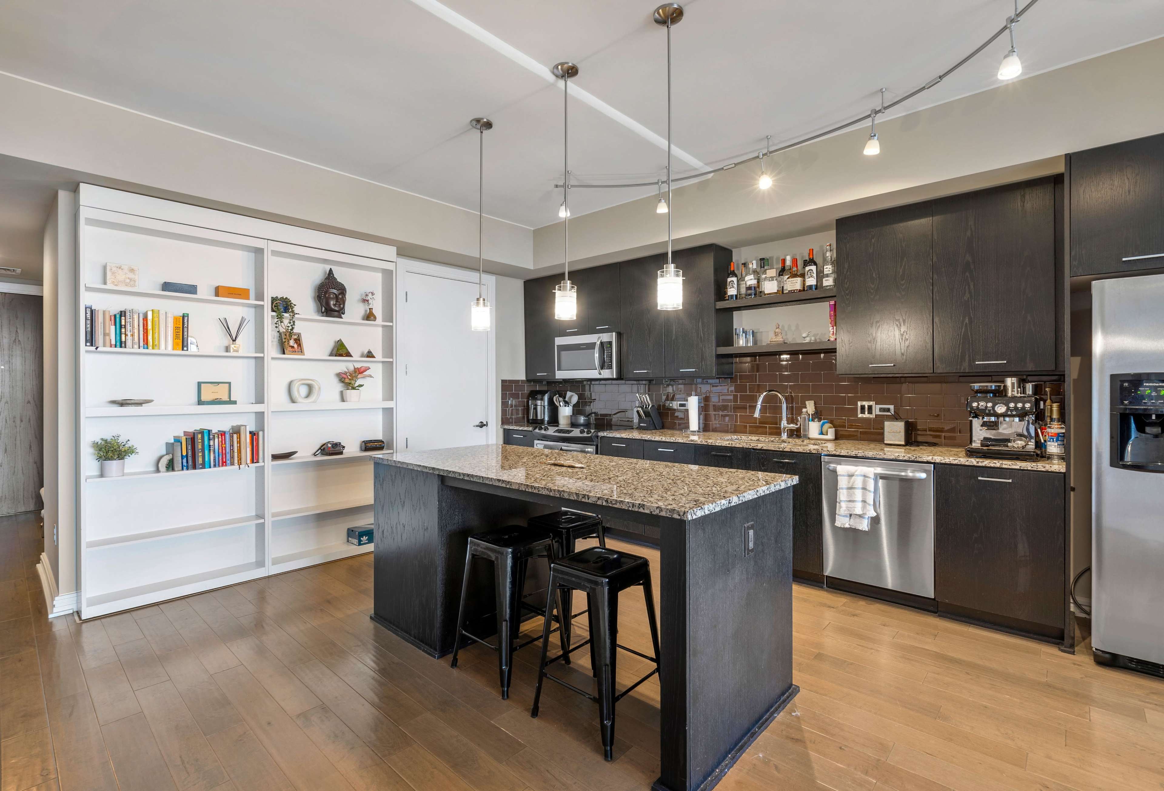 The image shows a modern kitchen with a central island, dark cabinetry, a built-in bookshelf, and stainless steel appliances.