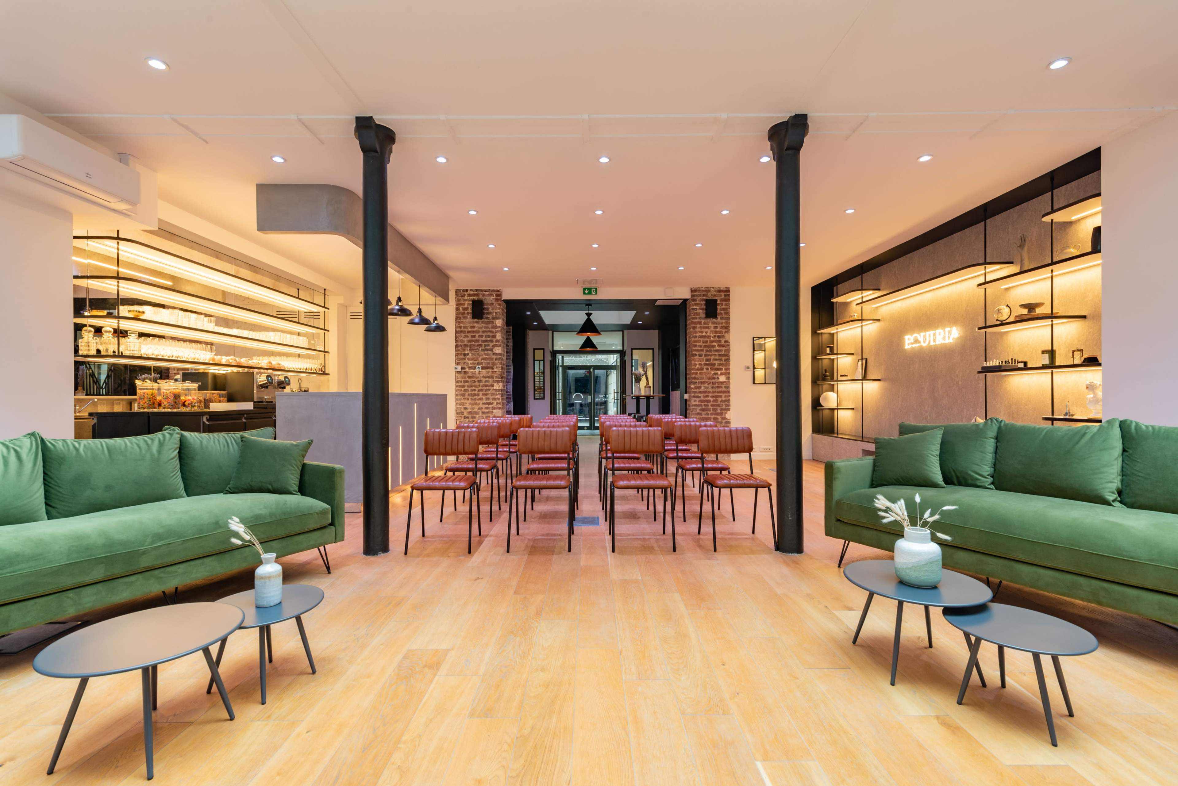 A modern lounge area with green sofas and small black tables, facing rows of seating in a well-lit space that leads to a bar and an entrance.