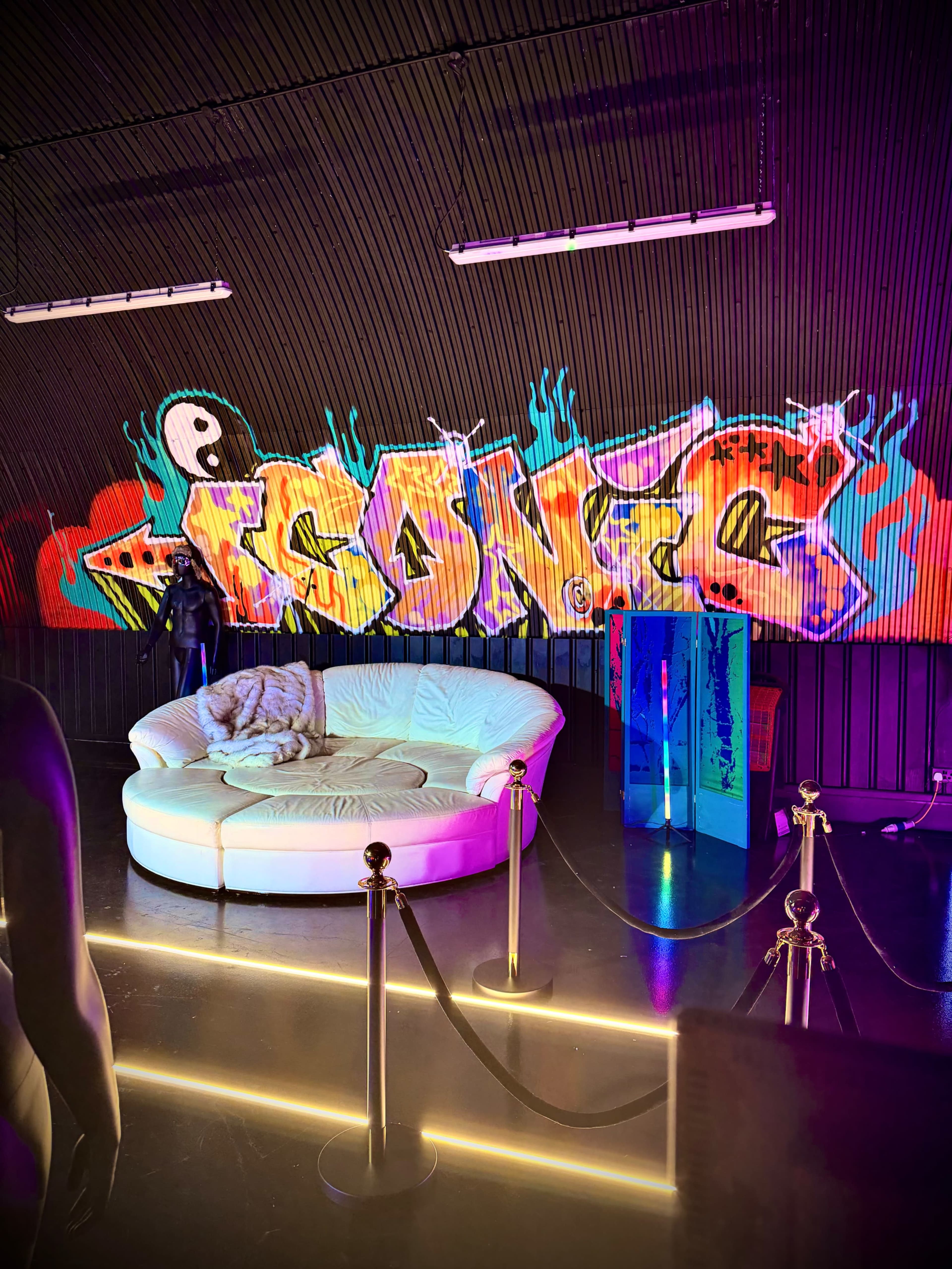 The image shows a brightly lit lounge area featuring a circular white sofa in front of a vibrant graffiti mural.