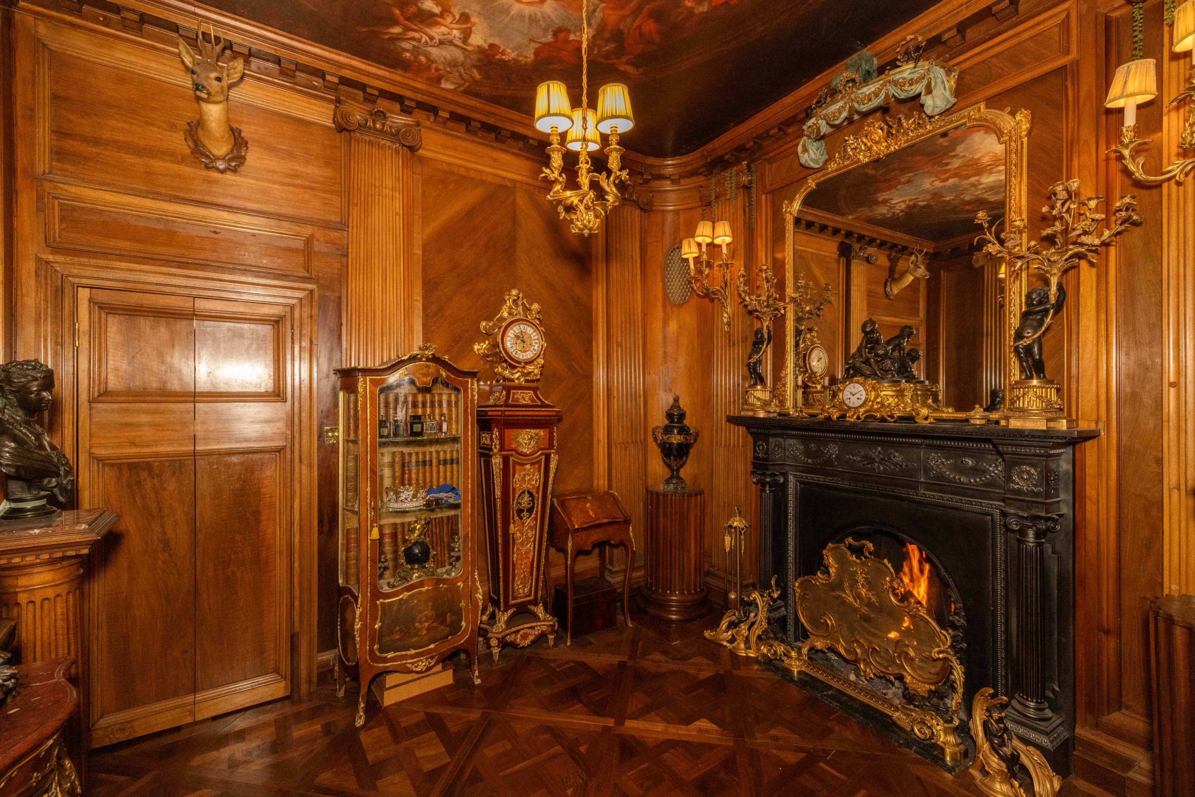 The room features ornate wooden paneling, a decorative fireplace, a vintage cabinet, and gilded details, illuminated by hanging lights.