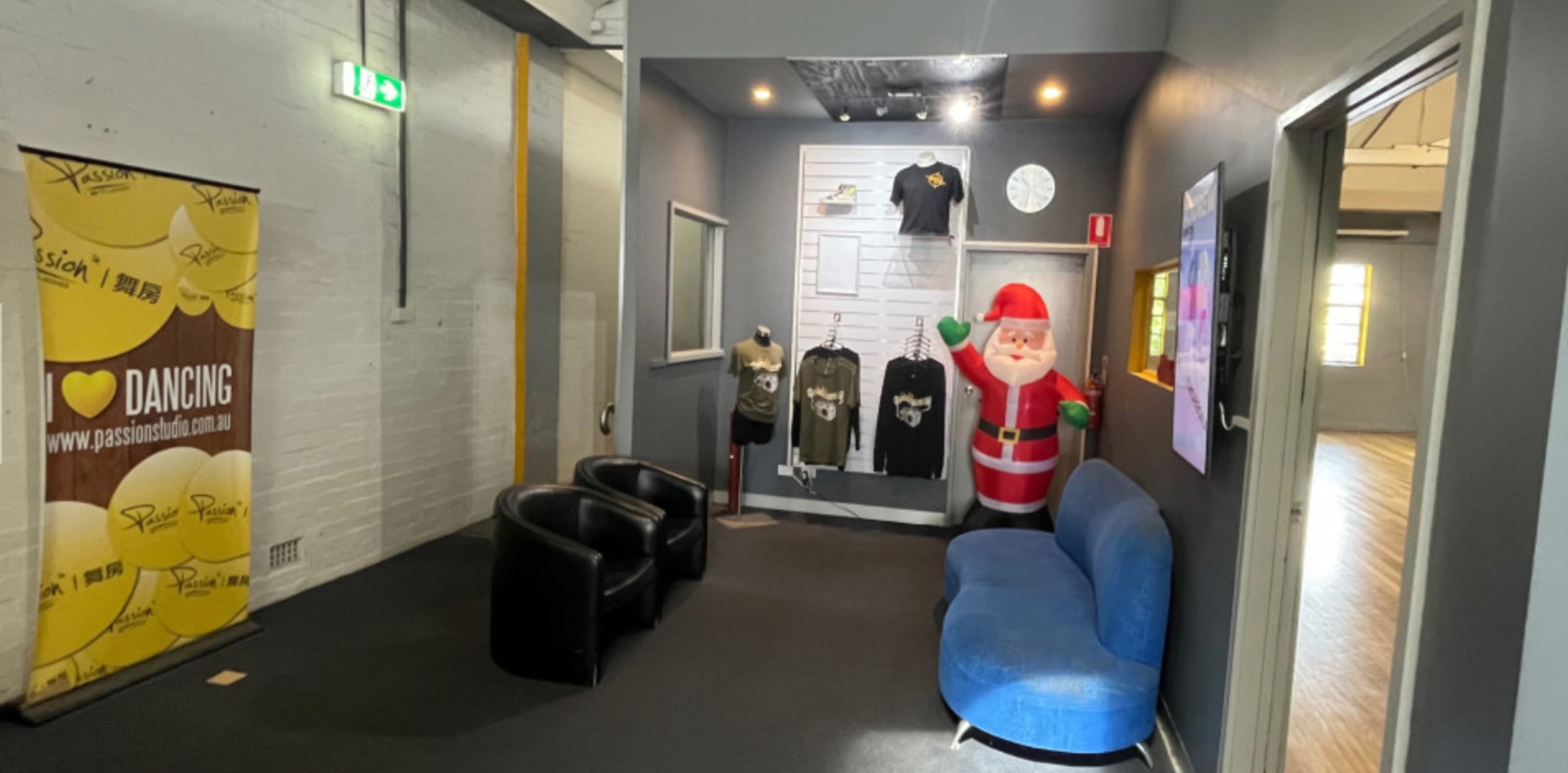 The image shows a waiting area featuring two black chairs, a blue couch, and a large inflatable Santa, with a wall displaying merchandise and a banner promoting dance.