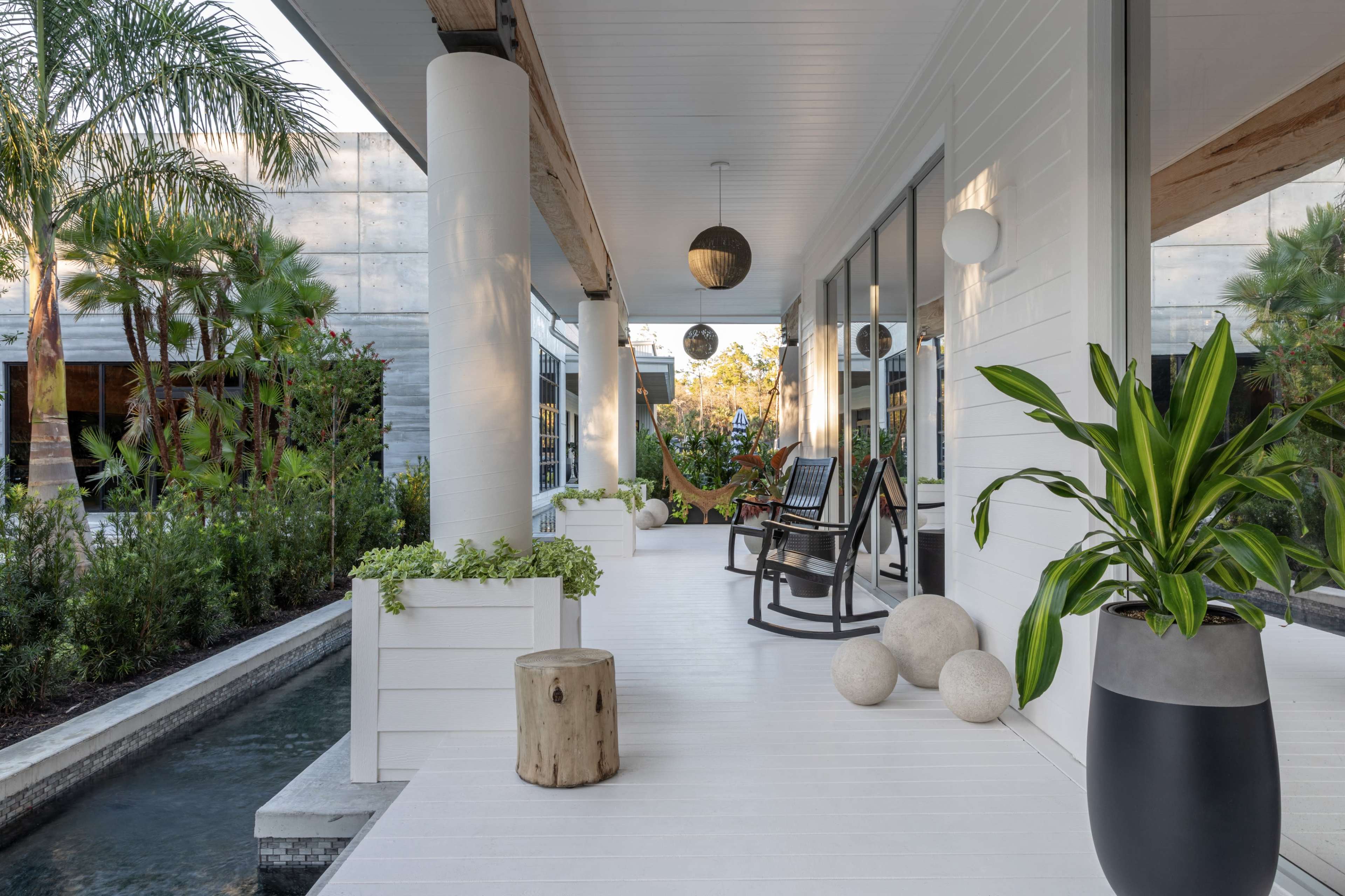 A spacious porch features rocking chairs, potted plants, and a hammock, surrounded by greenery and a water feature.