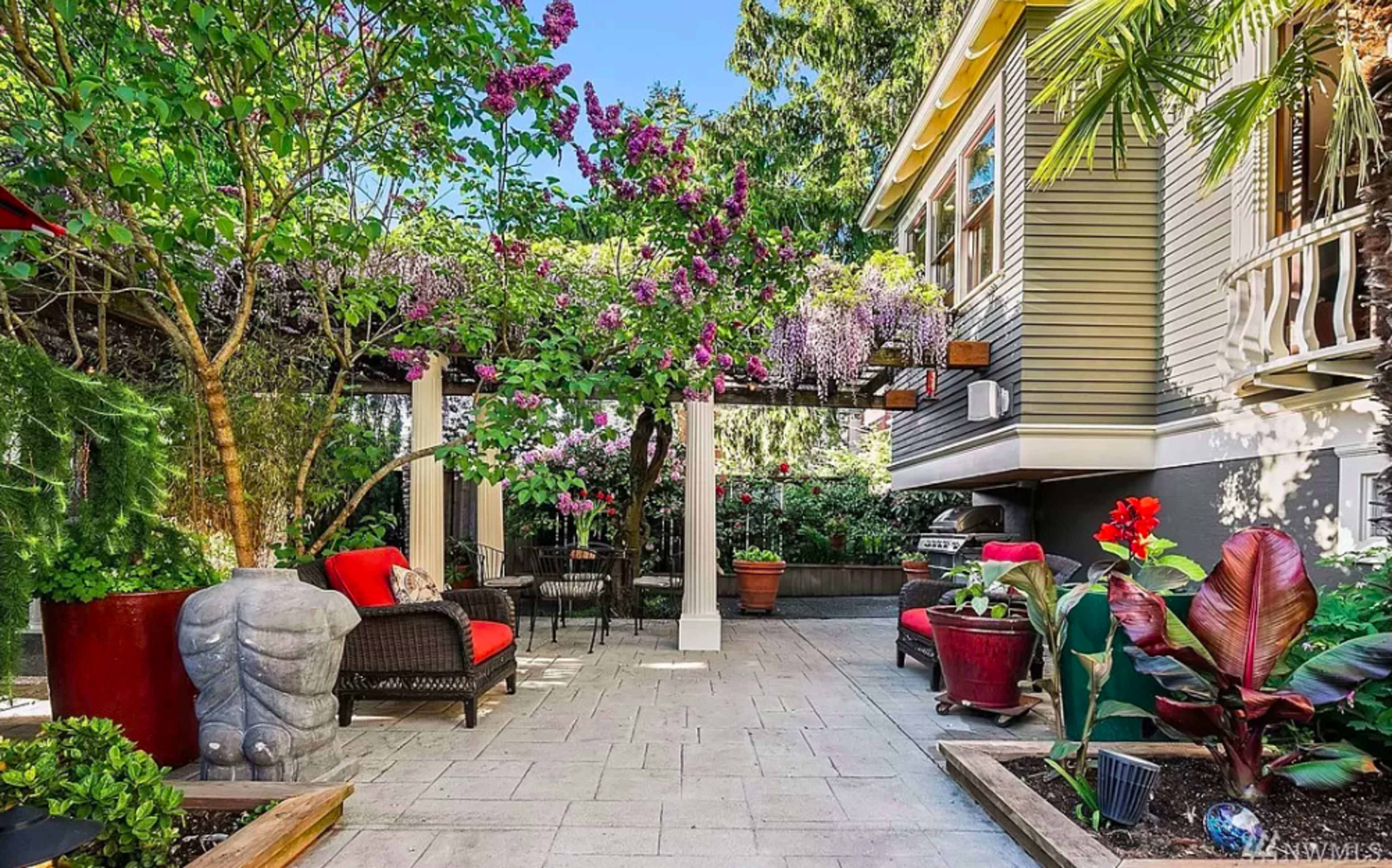 A landscaped outdoor patio features colorful plants and flowers, a Buddha statue, and seating arrangements near a house.
