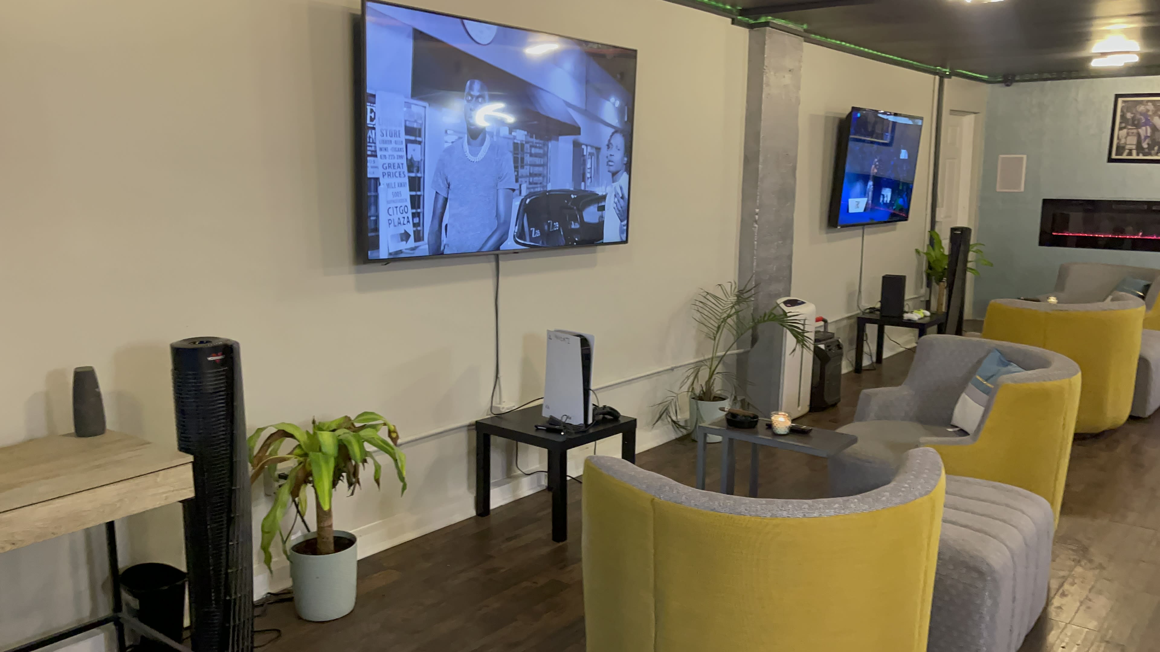 The image shows a cozy lounge area with two televisions mounted on the walls, a sofa set, and plants scattered throughout the space.