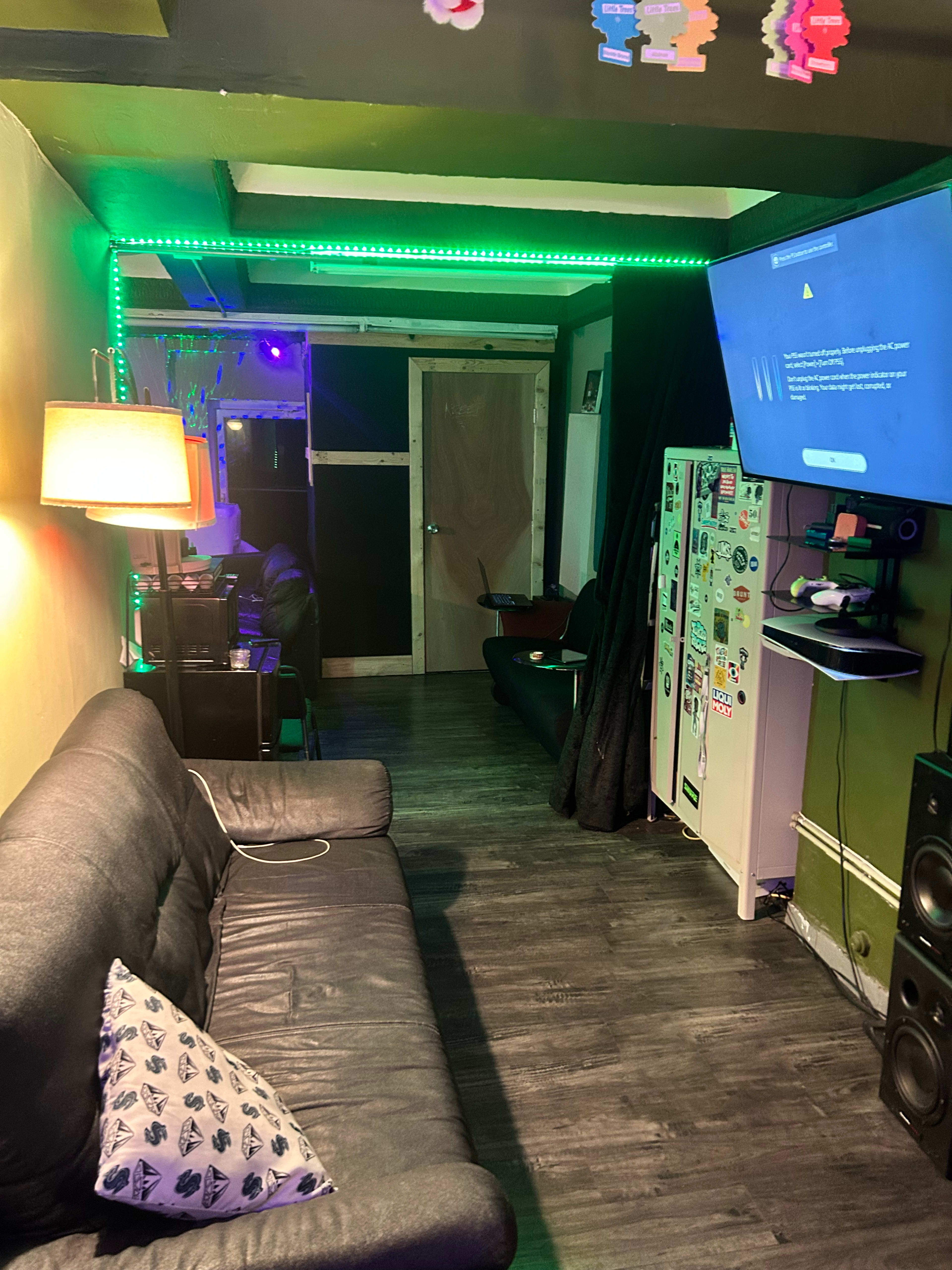 The image shows a cozy room with a black sofa, a television mounted on the wall, and green LED lighting along the ceiling.