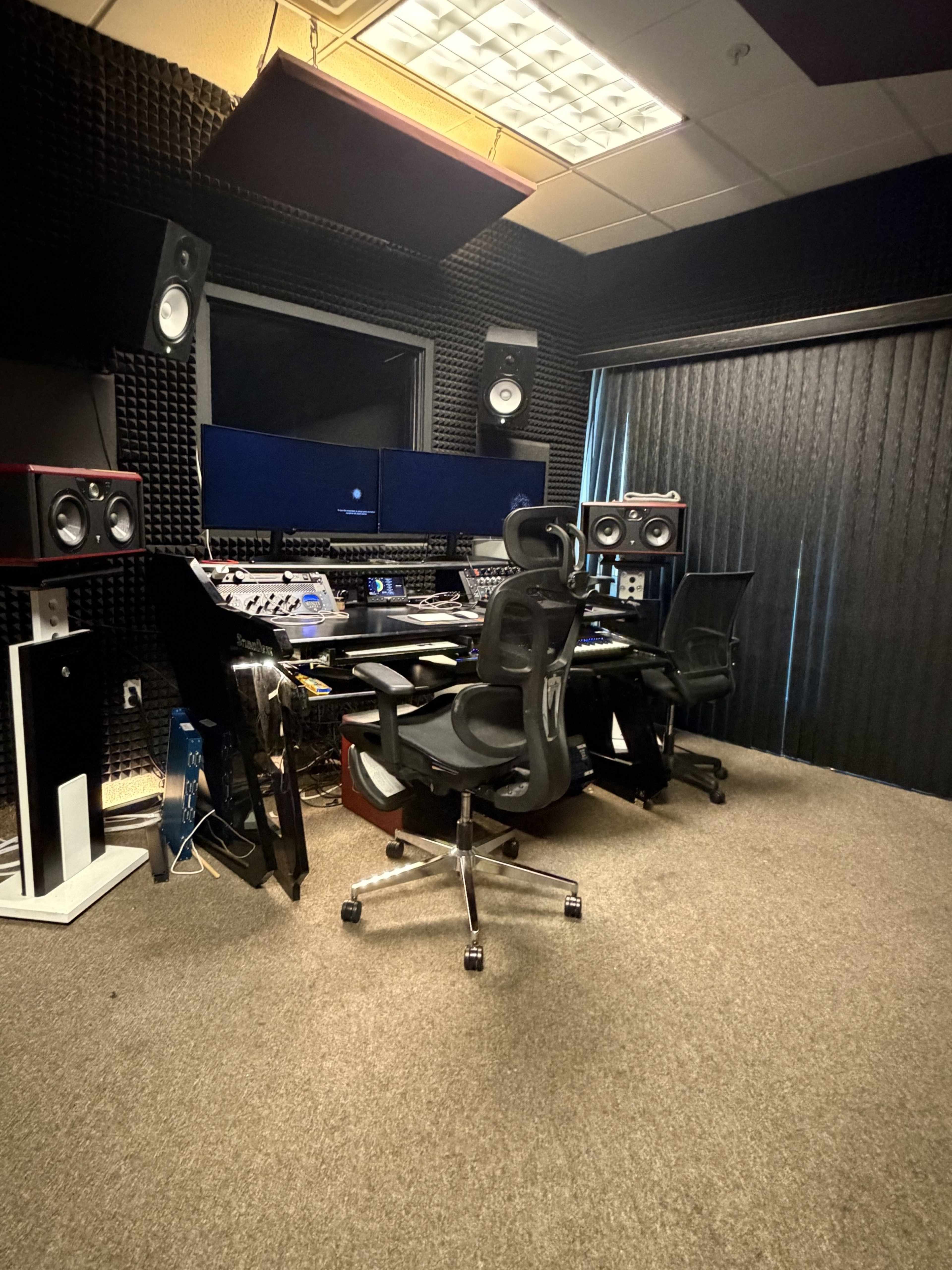 A sound studio is equipped with two monitors, audio speakers, and a central mixing console, surrounded by soundproofing panels.