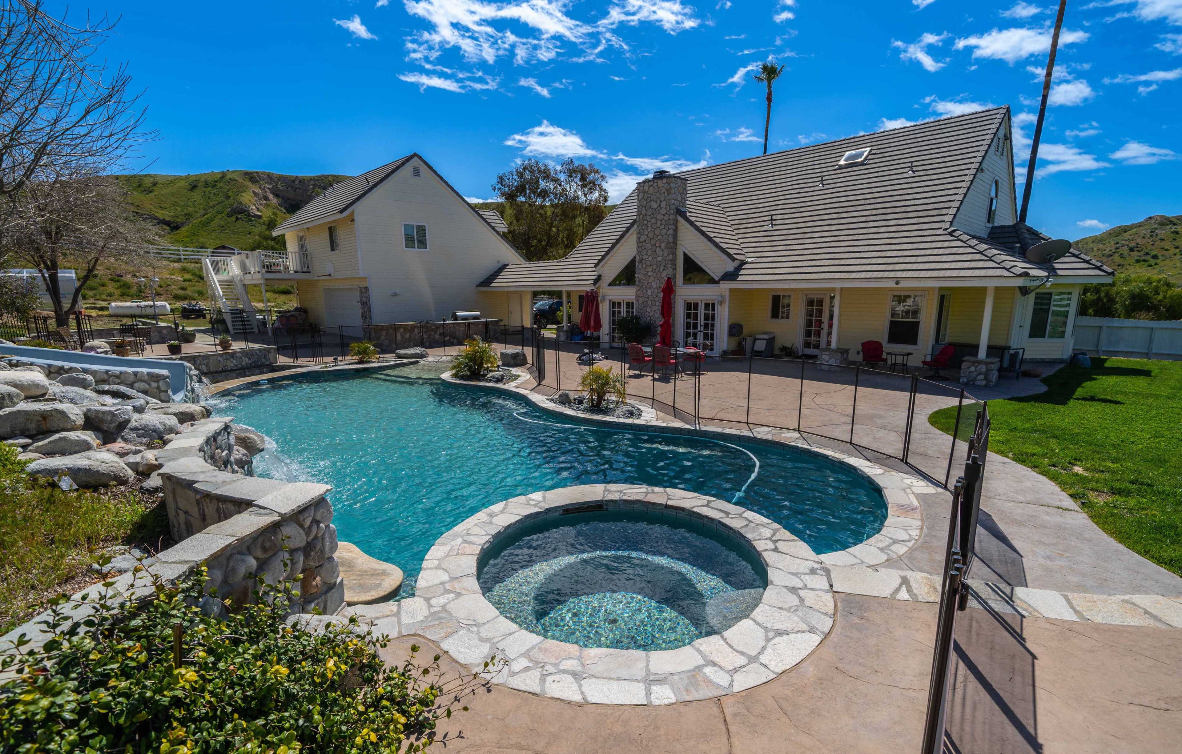 The image shows a backyard with a large swimming pool and a spa, surrounded by a stone patio and a house in the background.