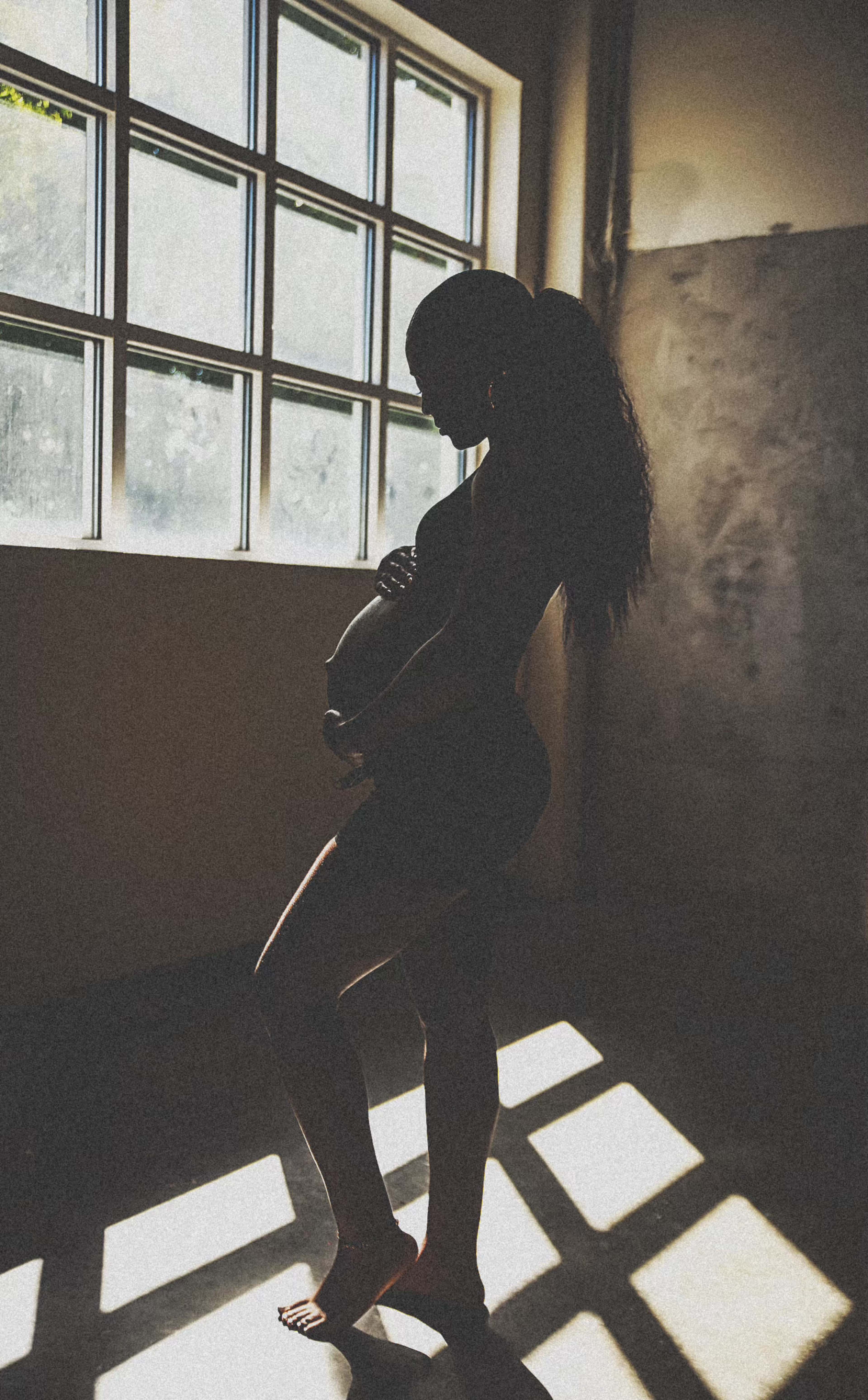A pregnant woman stands in silhouette, holding her belly near a window with sunlight casting geometric shadows on the floor.