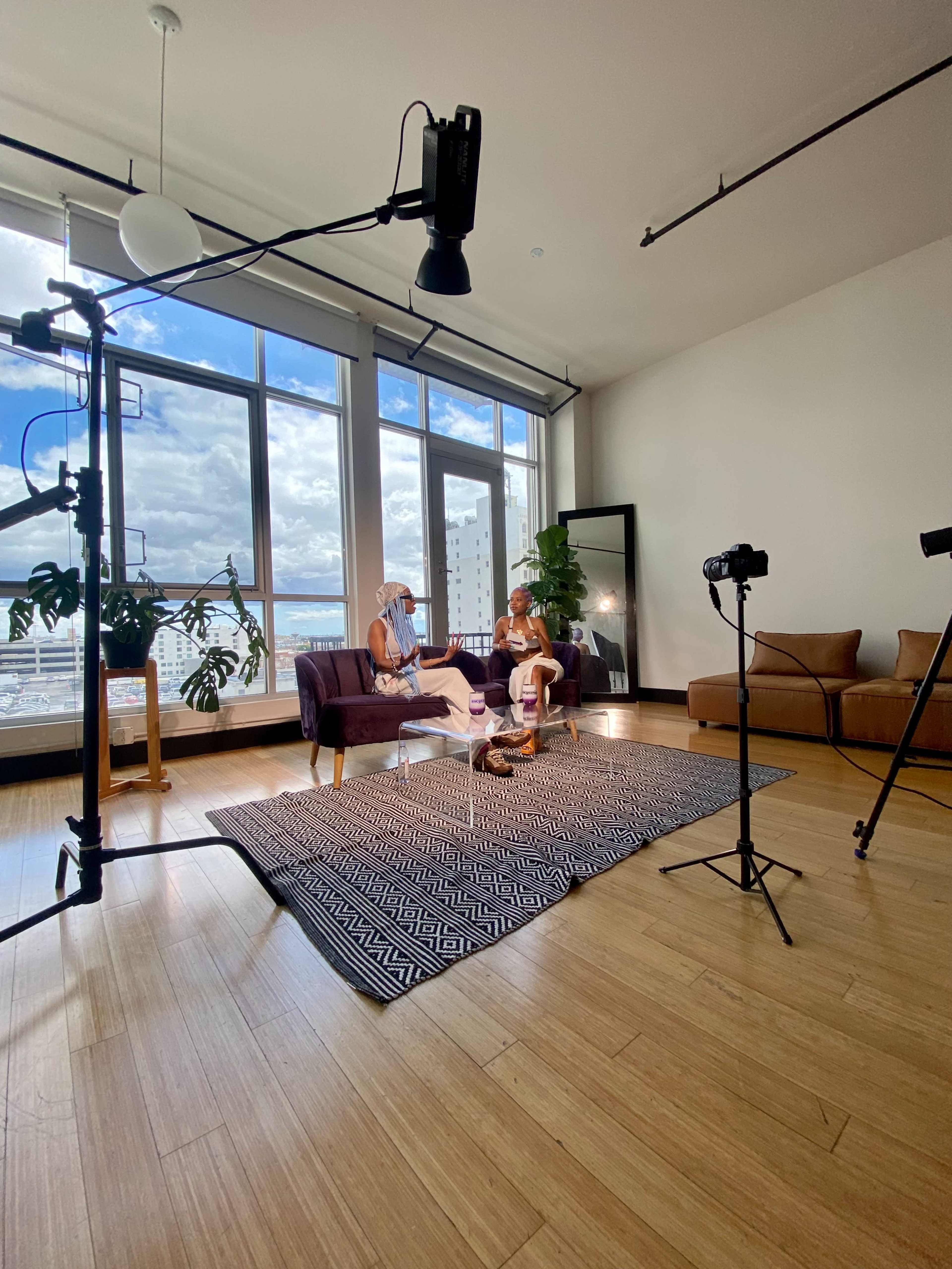 Two people sit on a purple couch in a brightly lit room with large windows, surrounded by filming equipment and plants.