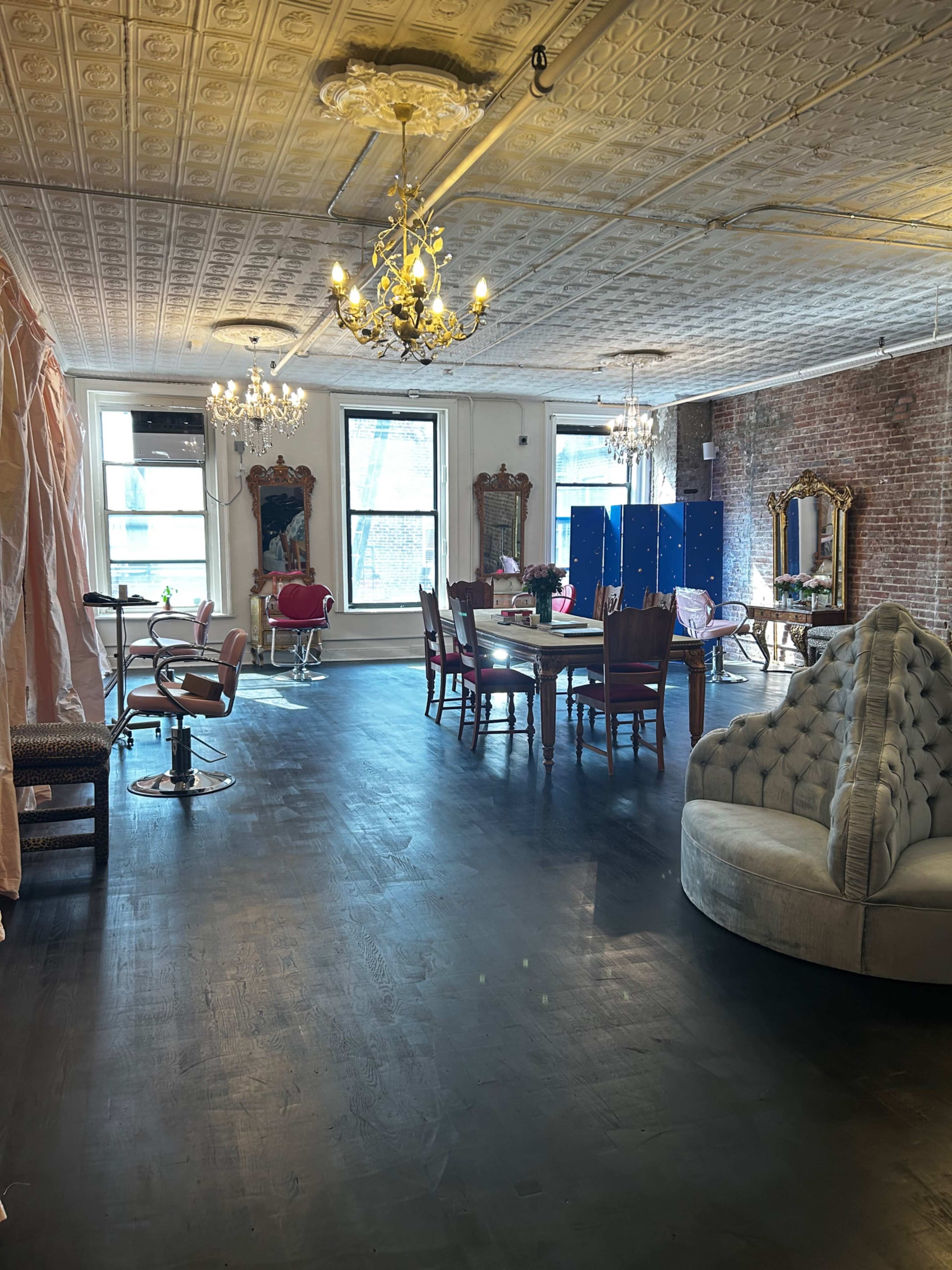 A spacious room with vintage-style furniture, chandeliers, and large windows, featuring a mixture of seating areas and a dining table.