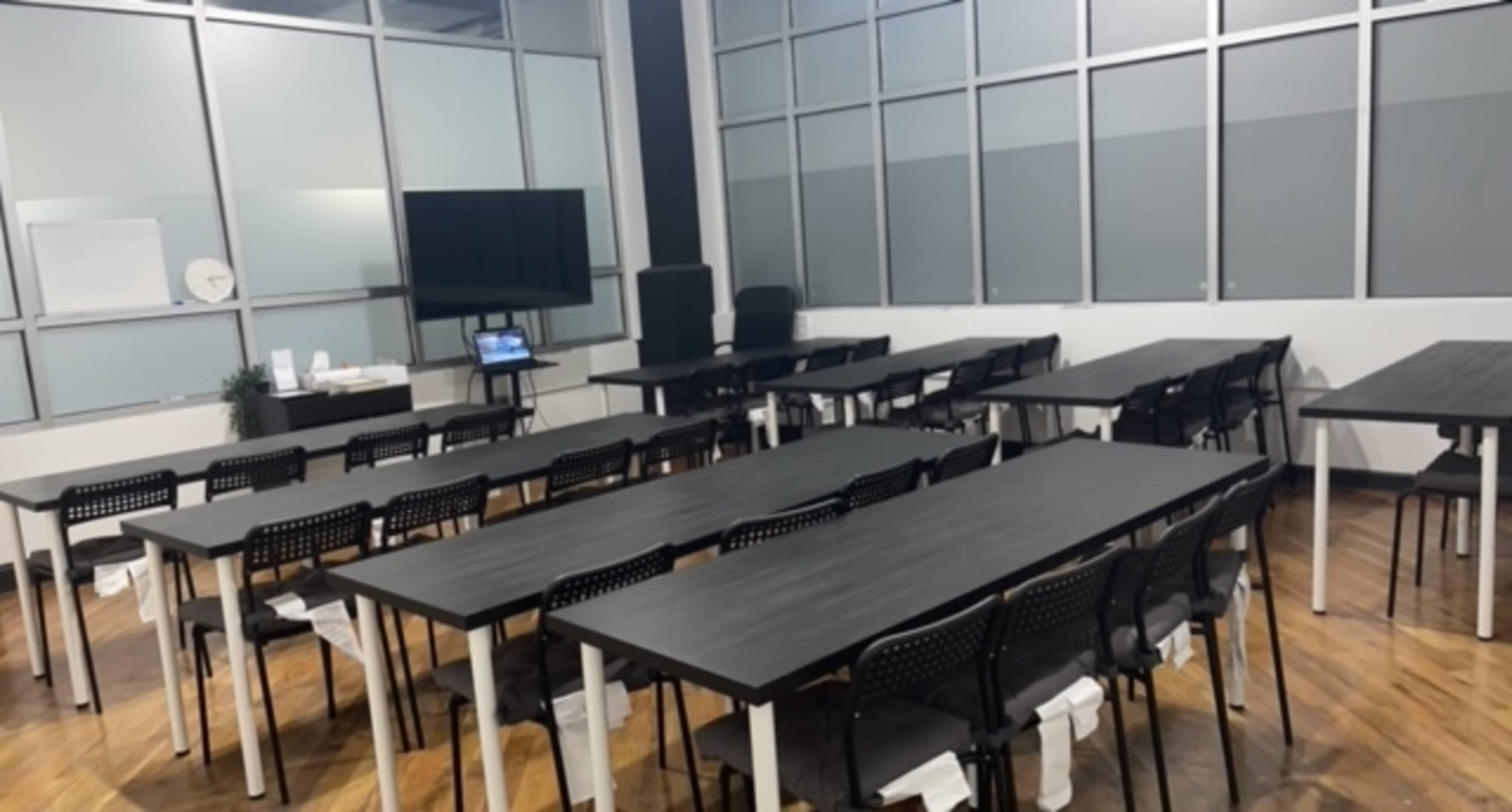 A spacious conference room is arranged with multiple long tables and chairs, accompanied by a television and a whiteboard.