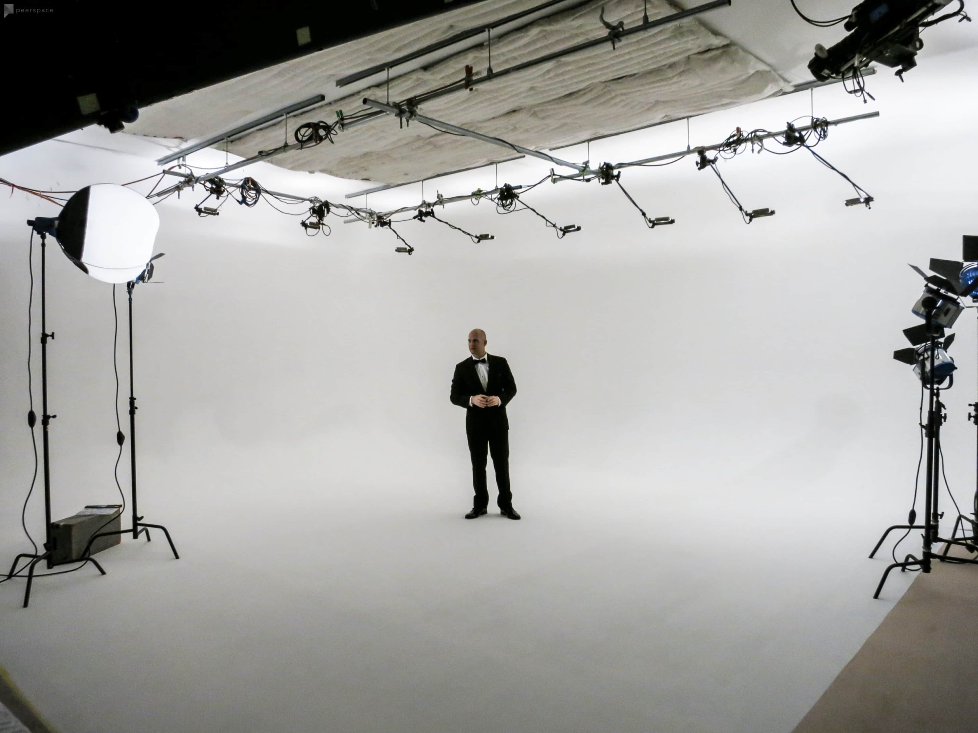 A man in formal attire stands alone in a sparsely populated photography studio with white walls and various lighting equipment.