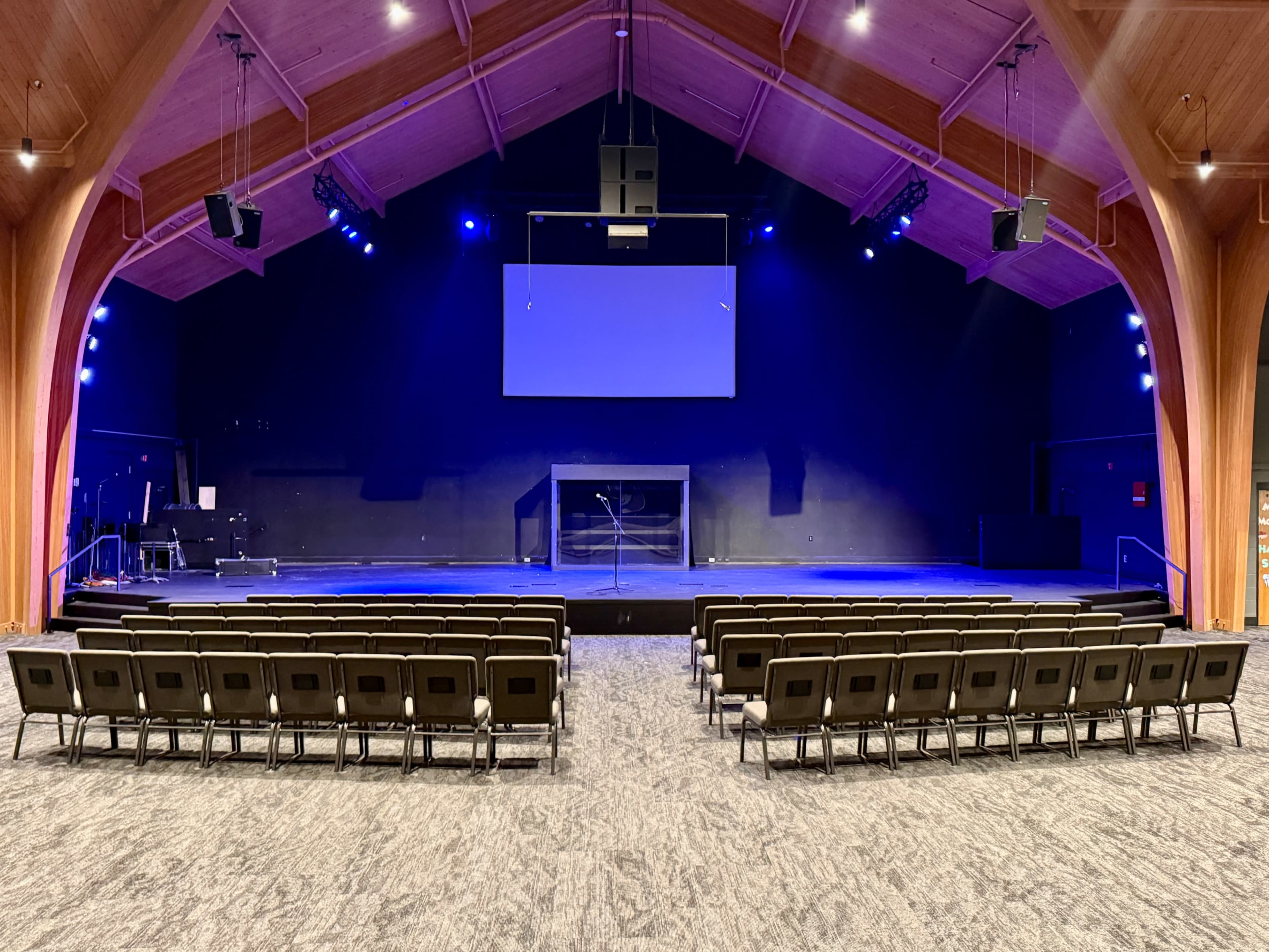 The image shows a stage area with a large, empty seating arrangement facing a backdrop and screen, illuminated by blue lights.