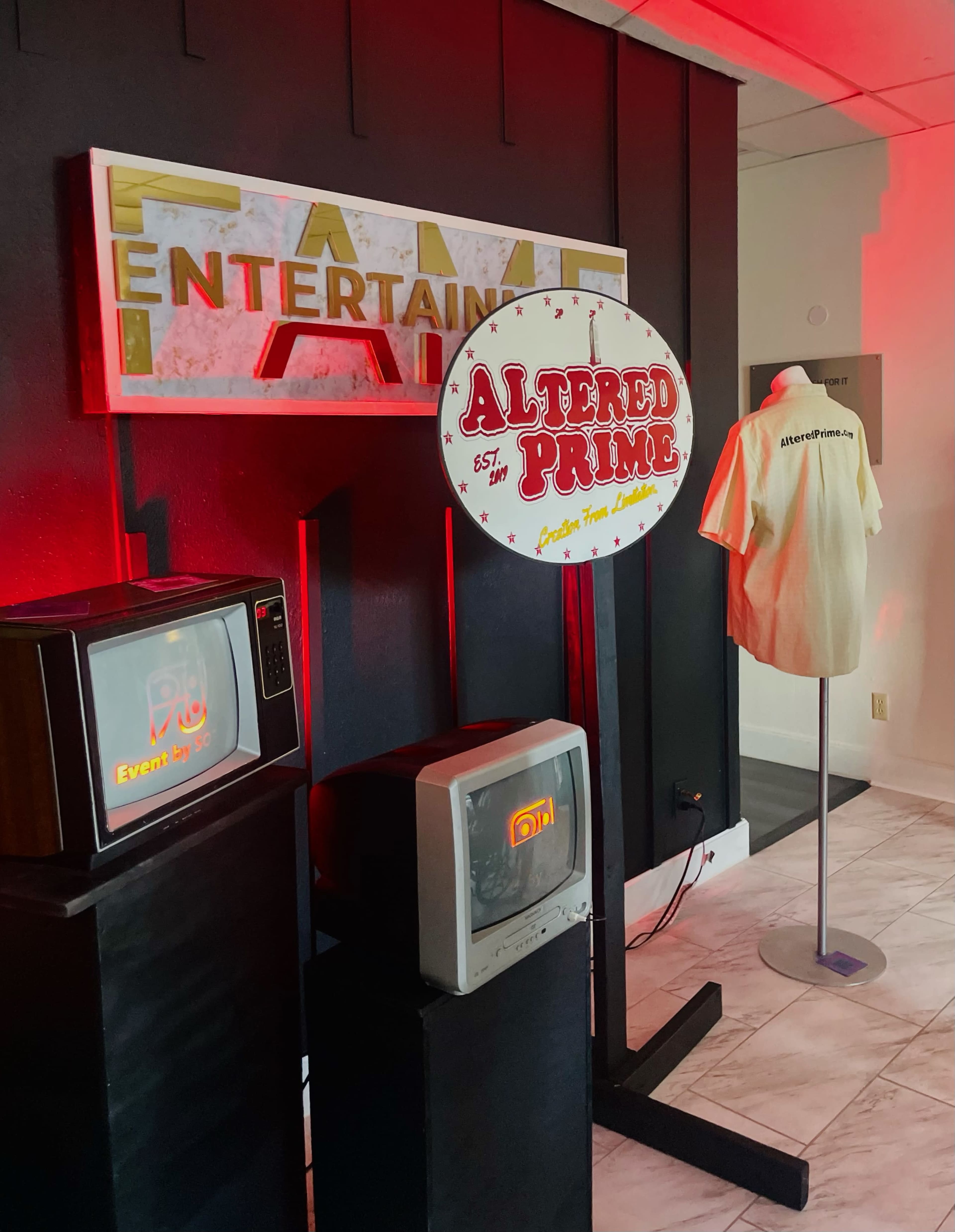 The image shows a display featuring a retro television, a vintage microwave, a sign for "Altered Prime" with a light-up design, and a shirt on a mannequin against a dark wall.