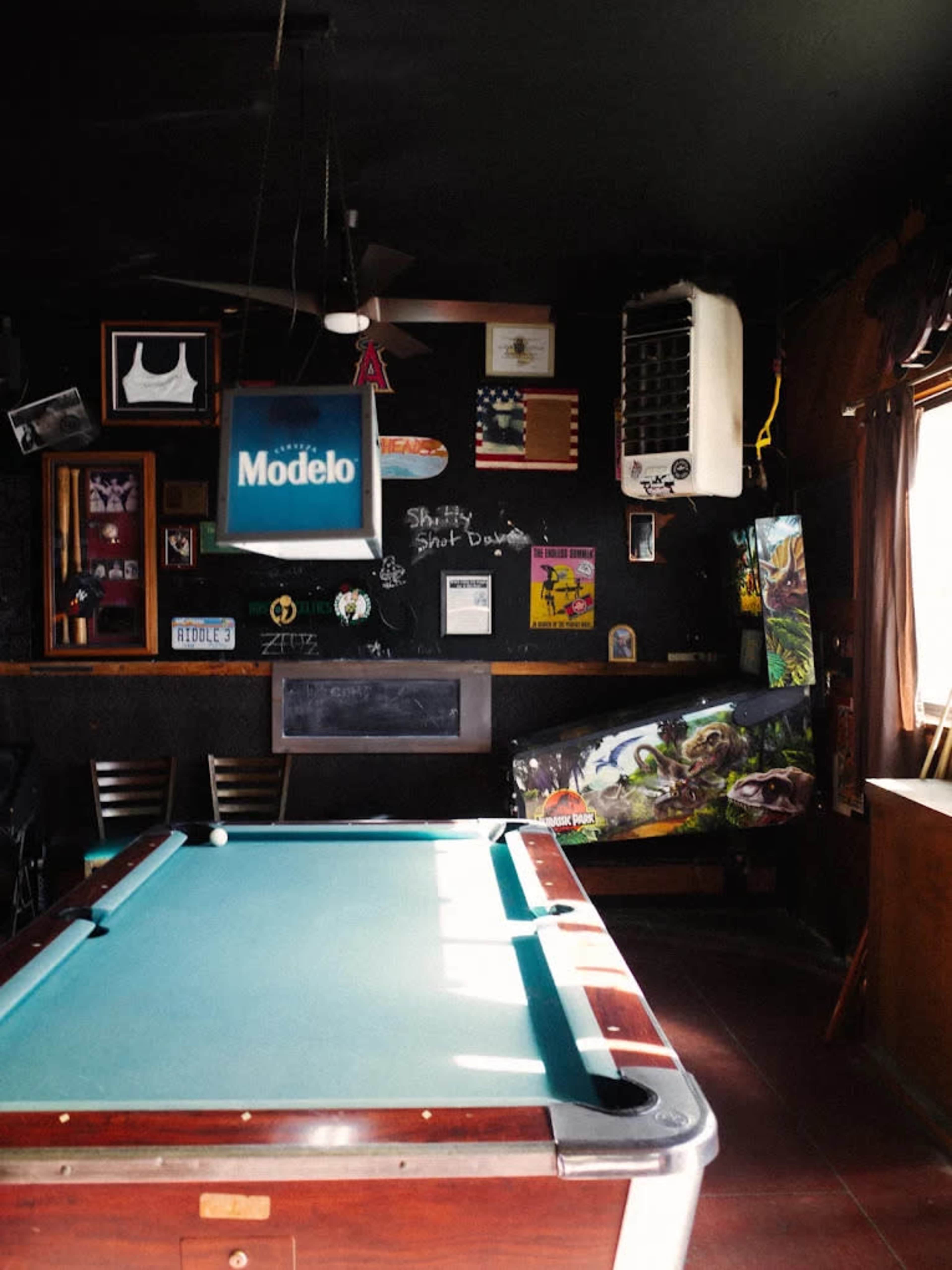 The image shows a dimly lit pool hall with a green felt pool table, decorated walls featuring various posters and memorabilia, and a window letting in natural light.