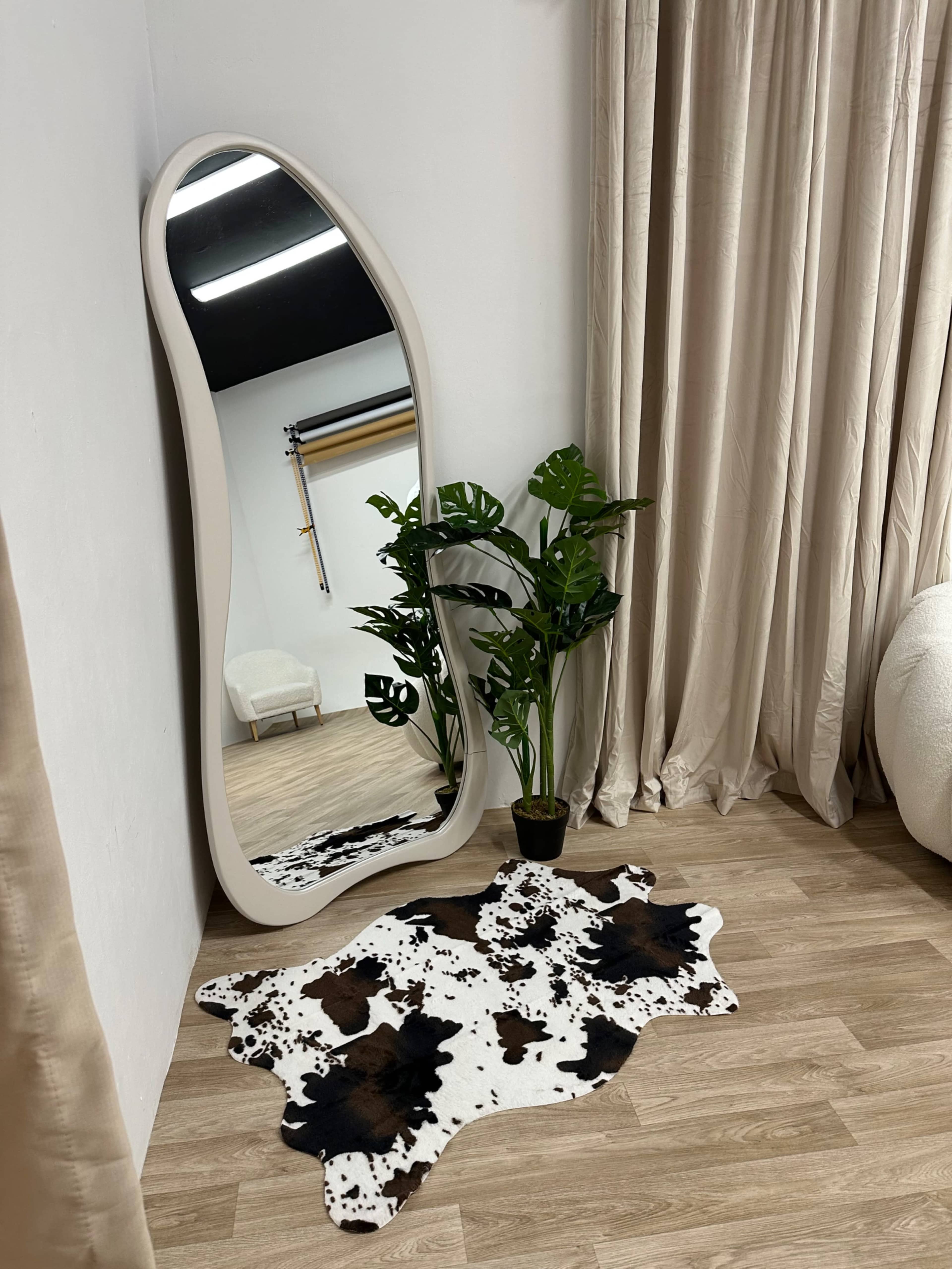 A full-length mirror with an organic shape stands near a potted plant on a cowhide rug, situated against a wall with beige curtains.